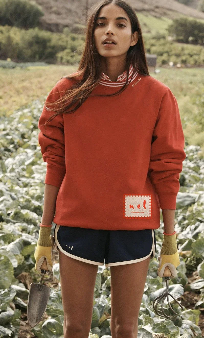A woman with long dark hair standing outdoors in a garden or farm, holding a small gardening tool in each hand, wearing a red sweatshirt, black shorts, and yellow gardening gloves.