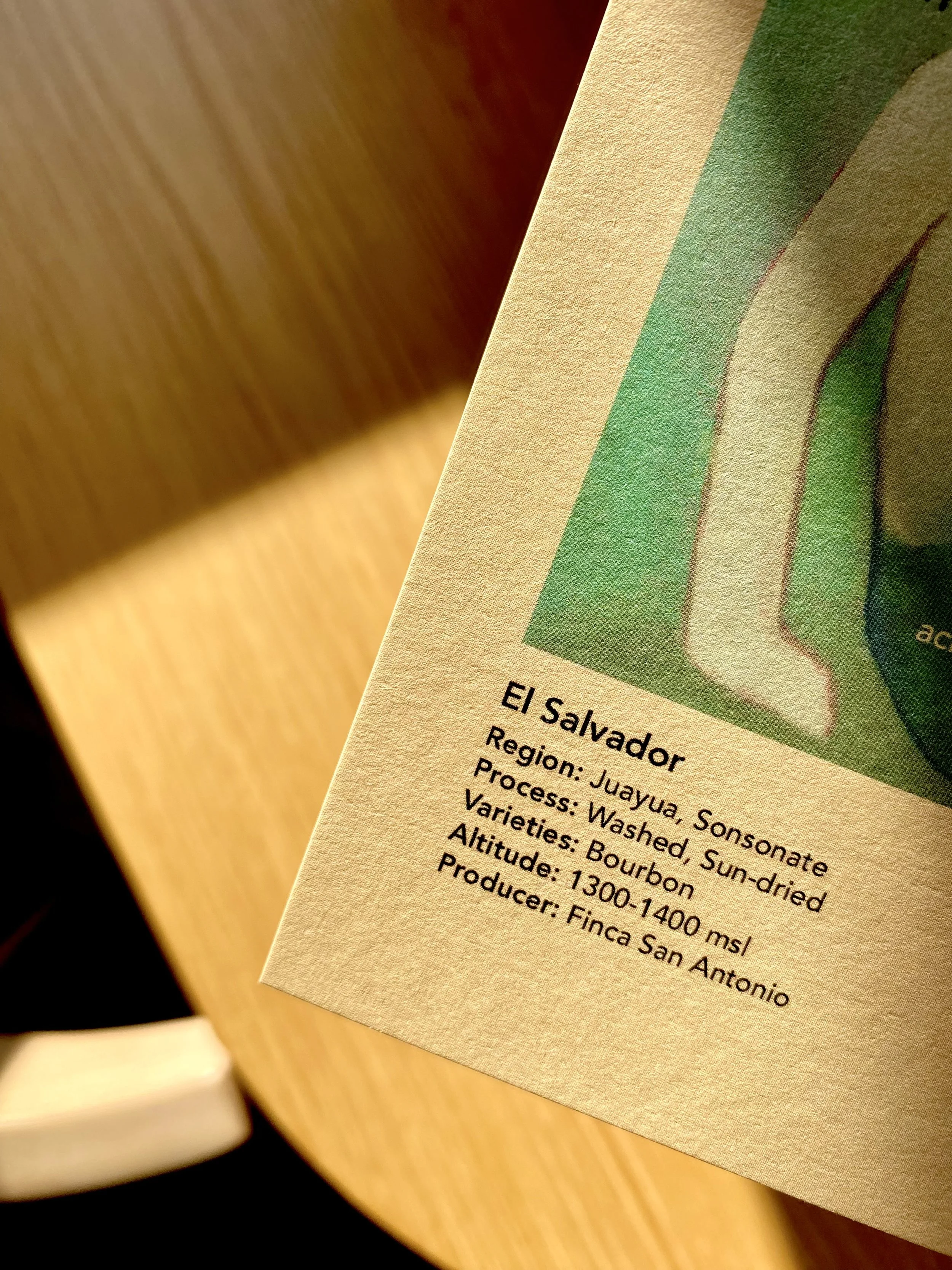 Close-up of a paper label on coffee packaging with details about the coffee, including origin from El Salvador, Juayua region, processed by washed, sun-dried method, bourbon variety, altitude of 1300-1400 meters, produced by Finca San Antonio.
