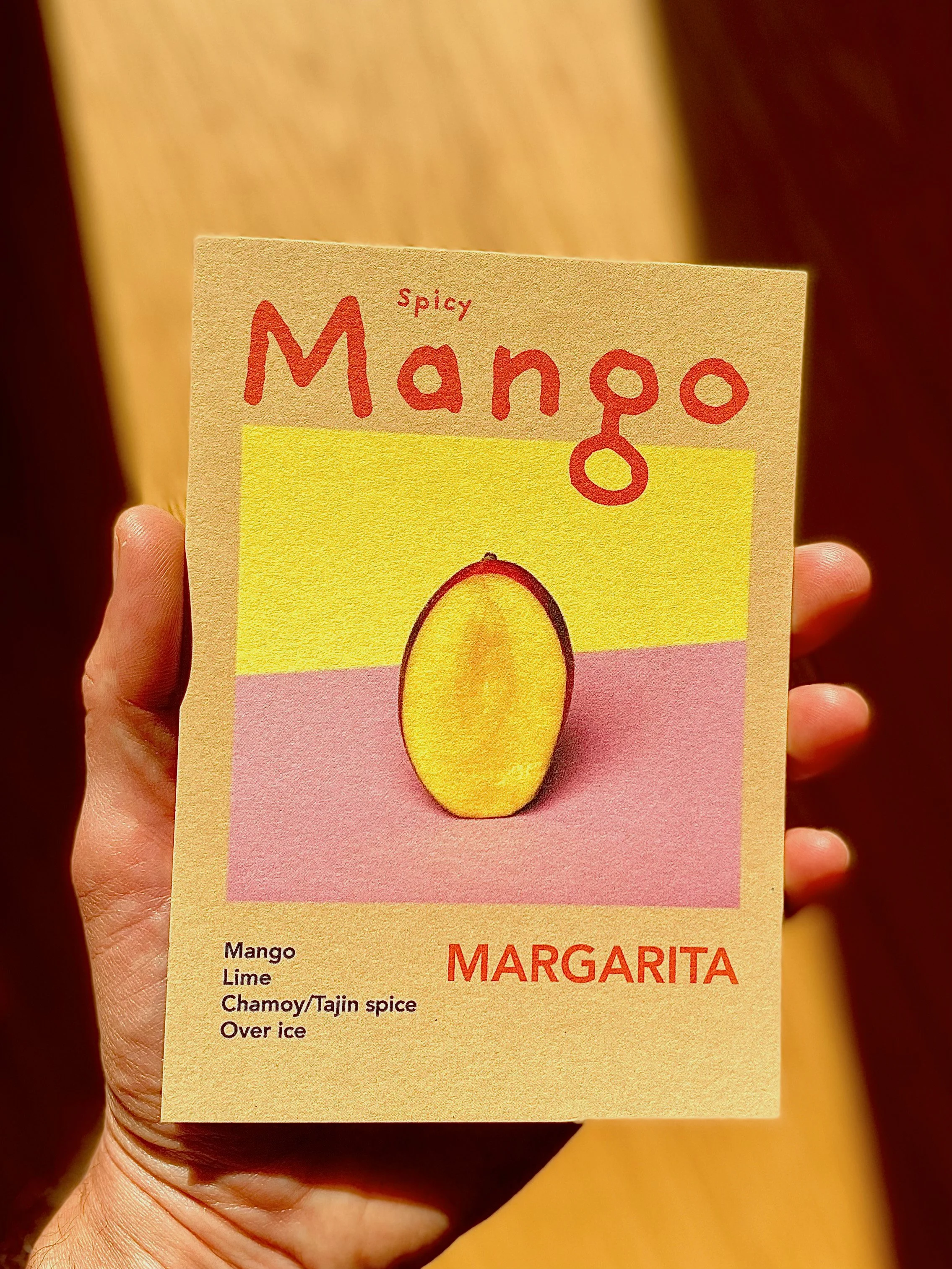 A hand holding a small, colorful paper menu featuring a mango margarita. The marketing menu has a yellow, pink, and beige background with an illustration of a mango.