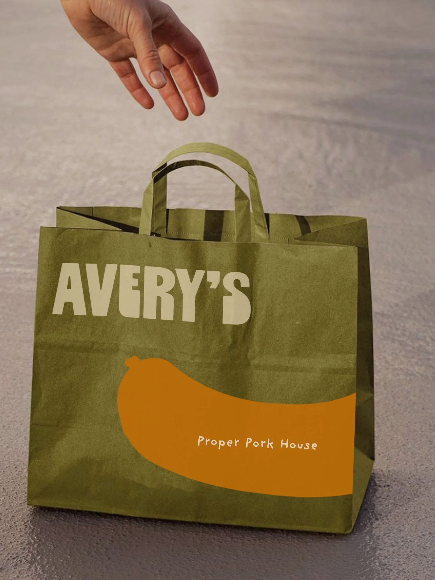 A green paper shopping bag with the brand name 'Avery's' in cream letters and a slogan 'Proper Pork House' in small white letters at the bottom and large mustard sausage logo. A hand is reaching down from the top, against a neutral background.