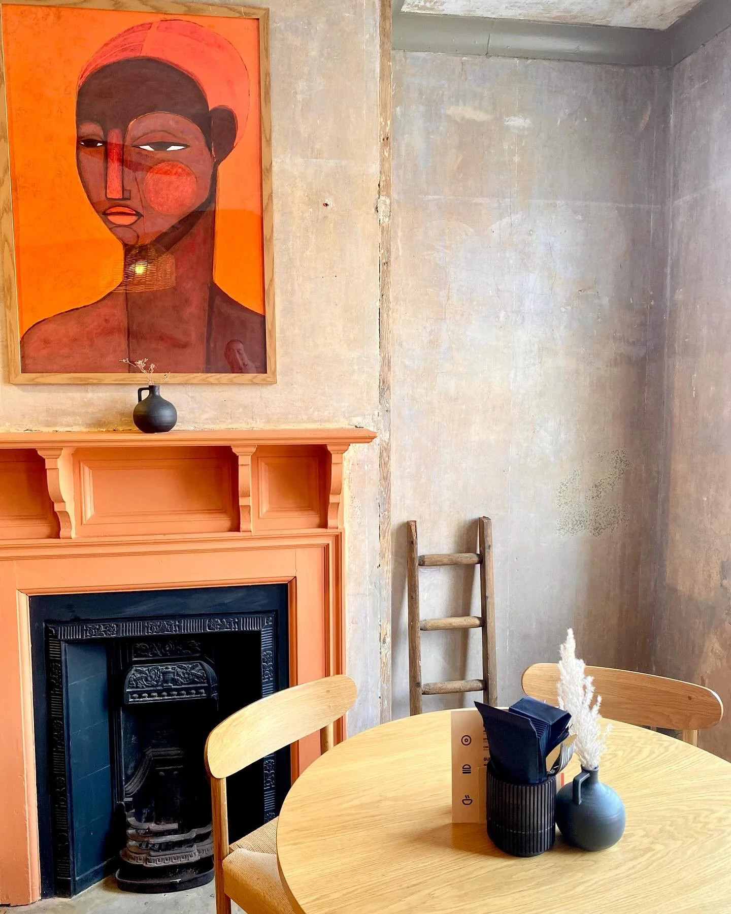 Interior of a rustic cafe or restaurant with a round wooden table, black vases with white decorative plants, wooden chairs, a wooden ladder, and a painting of a woman with dark skin, short hair, and a headscarf hanging on a textured wall above the fireplace.