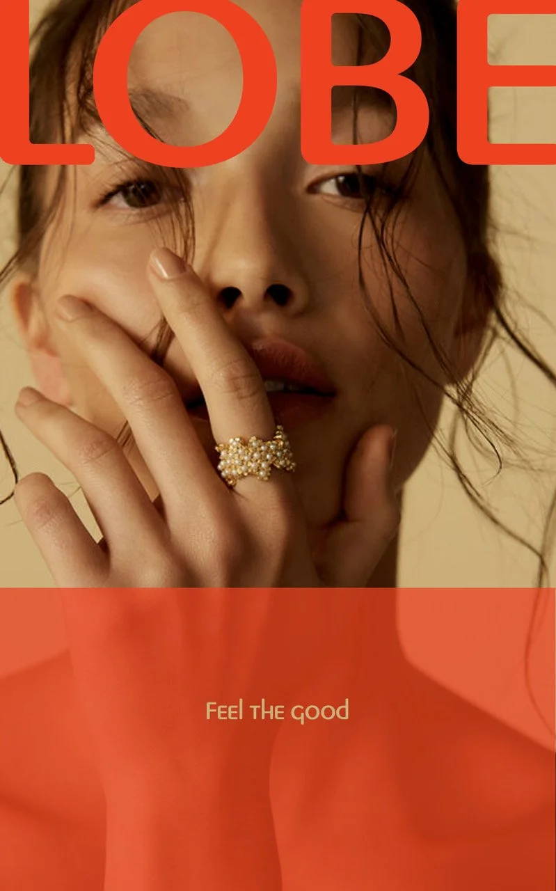 Close-up of a woman with dark hair and brown eyes, touching her face with her fingers. She is wearing a pearl ring and has natural makeup. Overlaid text reads 'LOBE' at the top and 'Feel the good' at the bottom.