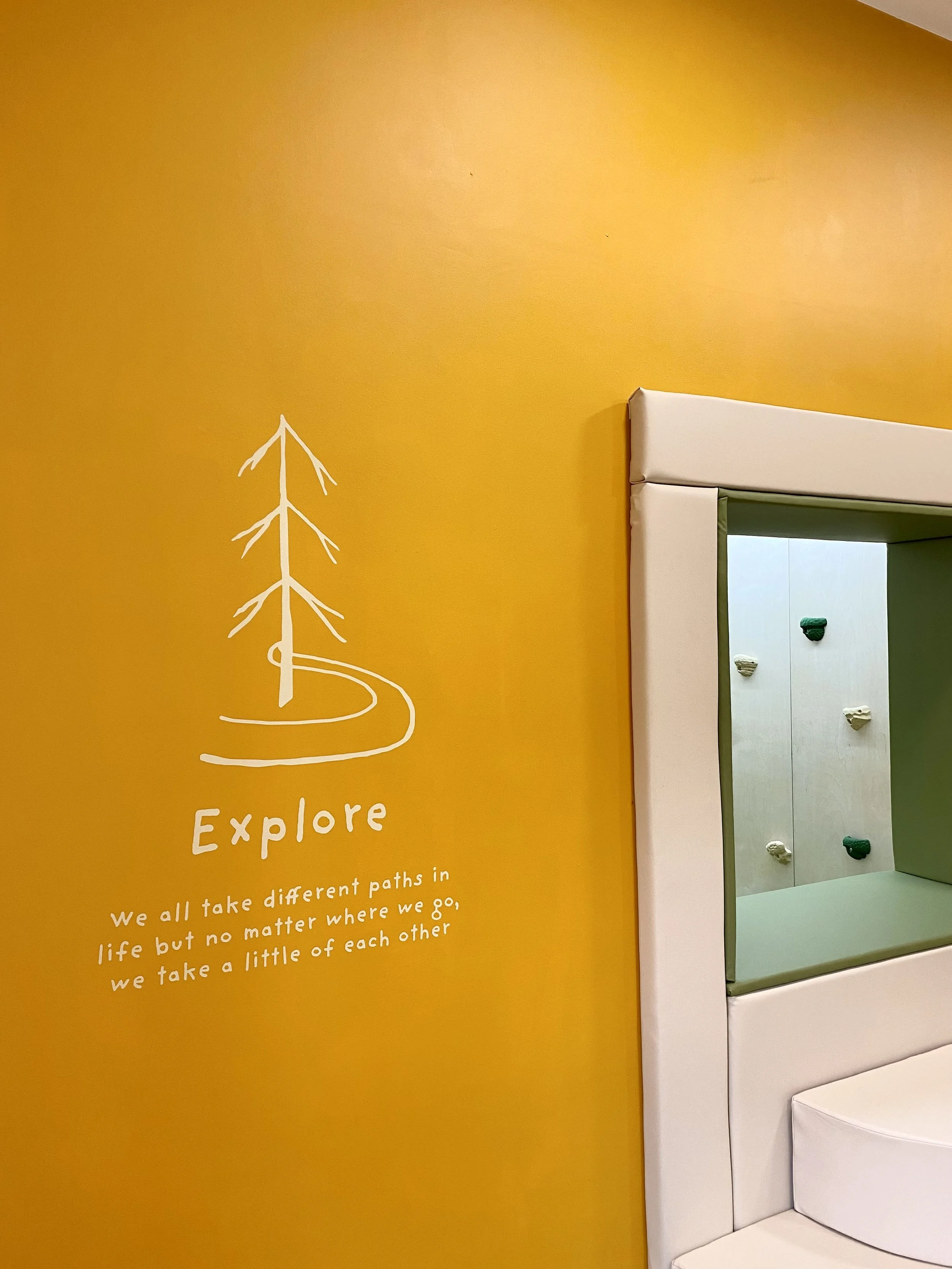 A yellow wall with ''EXPLORE' wall graphic, next to a white cushioned children's indoor climbing zone with climbing holds visible in the background.