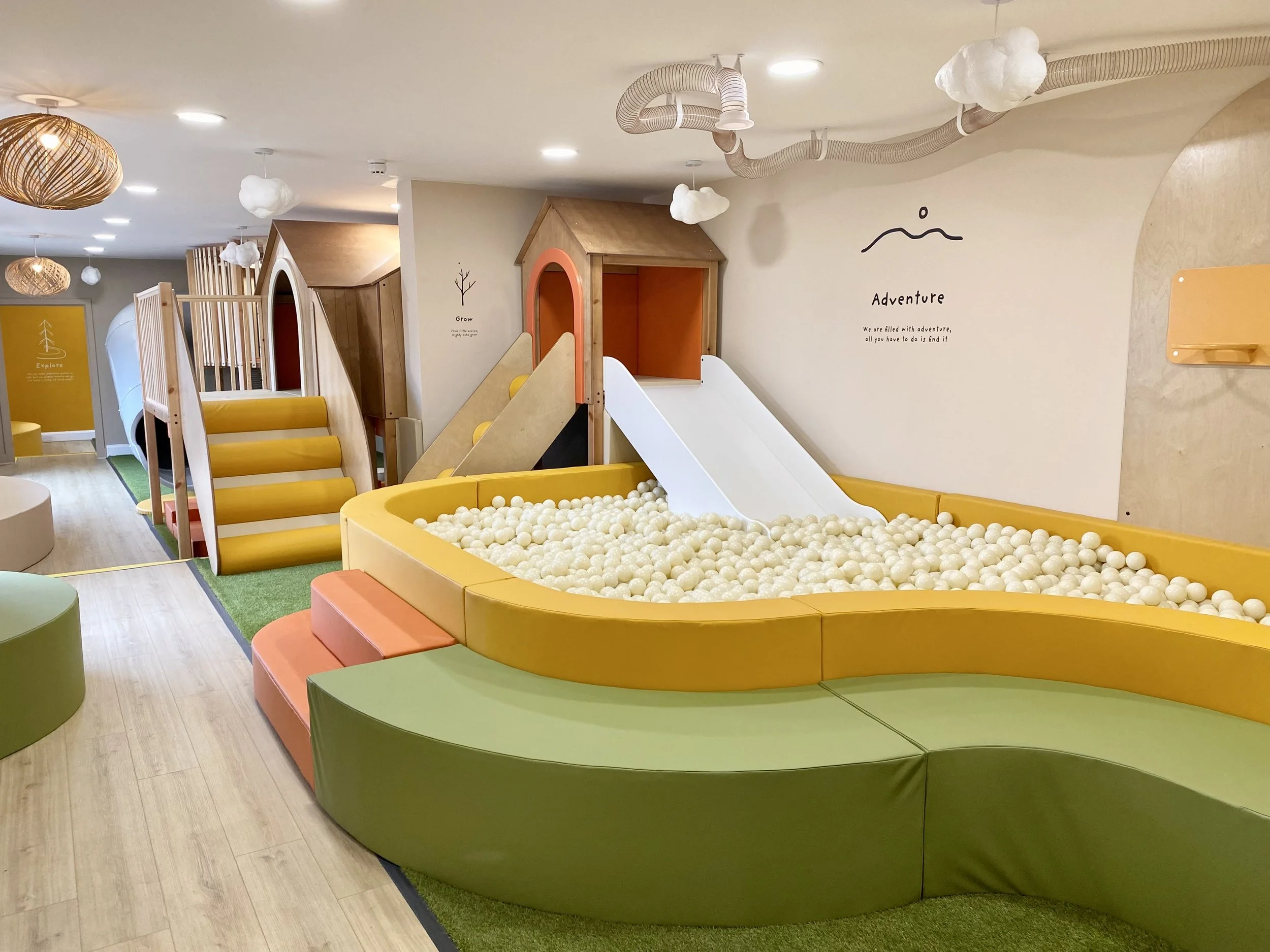 Indoor children's play area with soft, colorful cushioned seating, a ball pit, slides, and climbing structures, surrounded by wooden accents and decorative cloud-shaped lights.