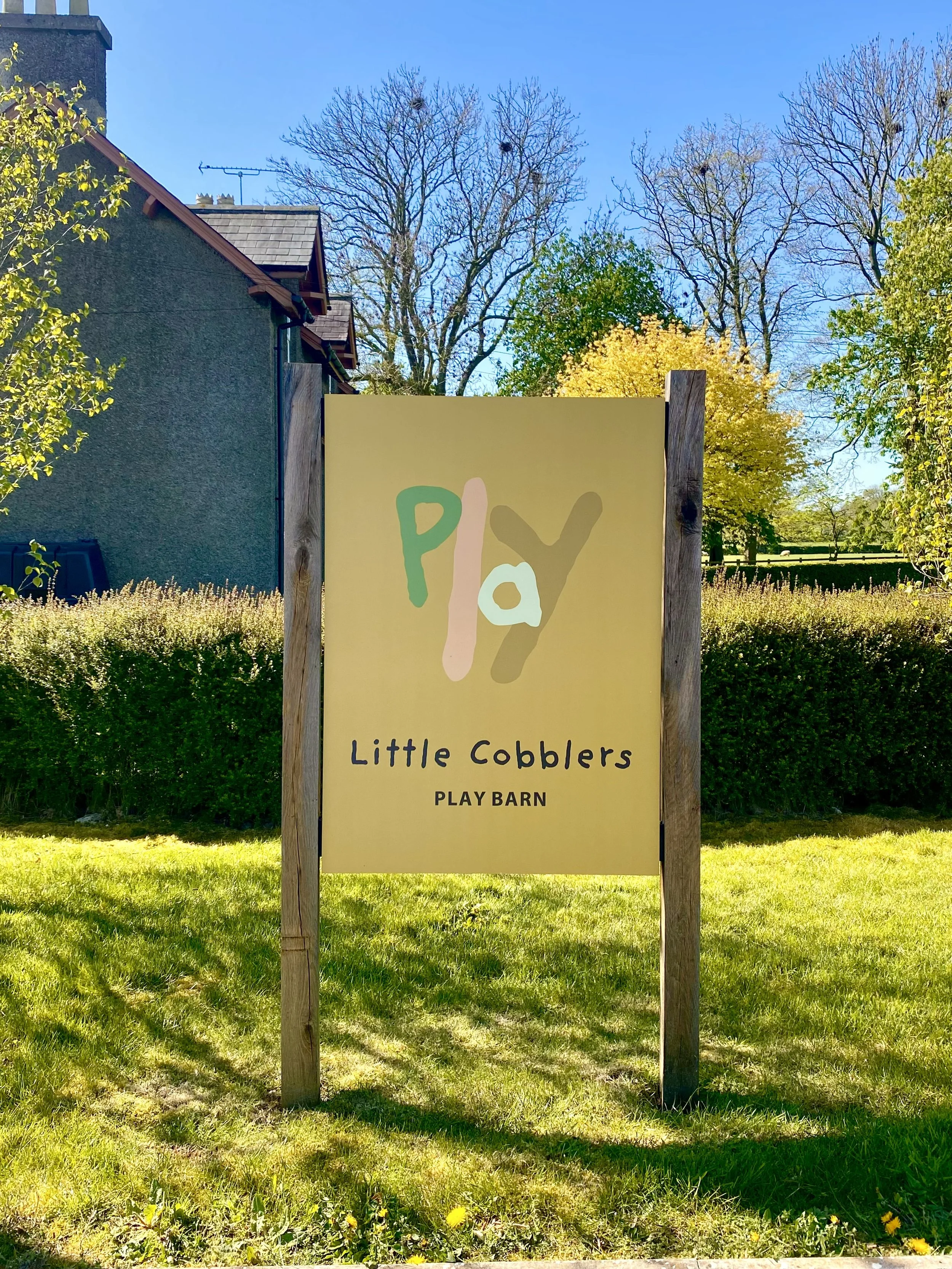 Sign that reads 'play' and 'Little Cobblers Play Barn' in front of a grassy area with trees, some with yellow and green leaves, under a clear blue sky.