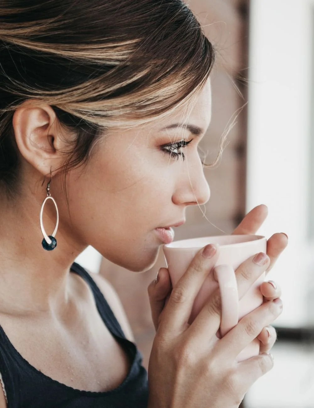A woman with light brown hair and dangling earrings is holding a white mug close to her lips, appearing to smell or drink from it.