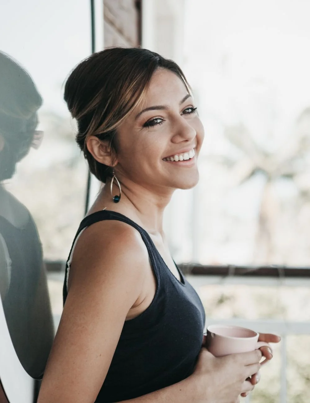 A smiling woman with short brown hair holding a pink mug, wearing earrings, and standing indoors near a large window with a blurred outdoor background.