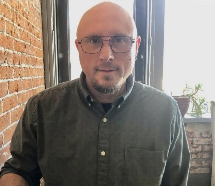 A bald man with glasses and a short beard wearing a button-up shirt, sitting indoors near a window and brick wall.