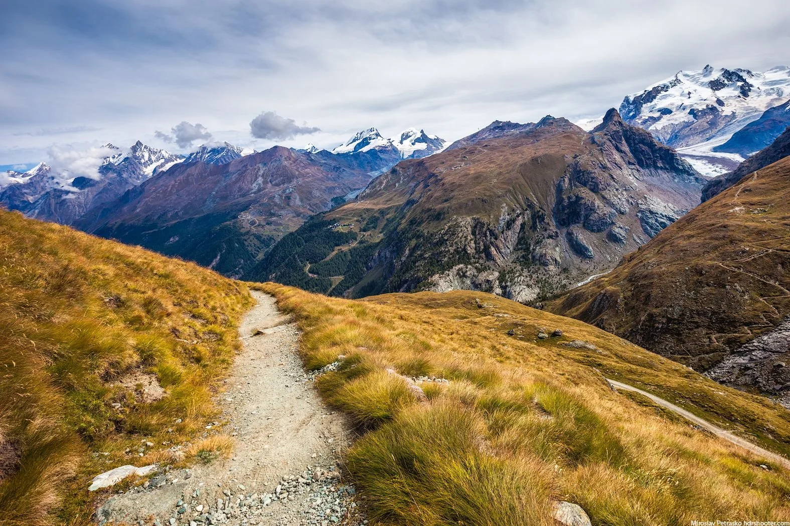 Mountain trail winding through grassy hills with snow-capped peaks in the distance under a cloudy sky.