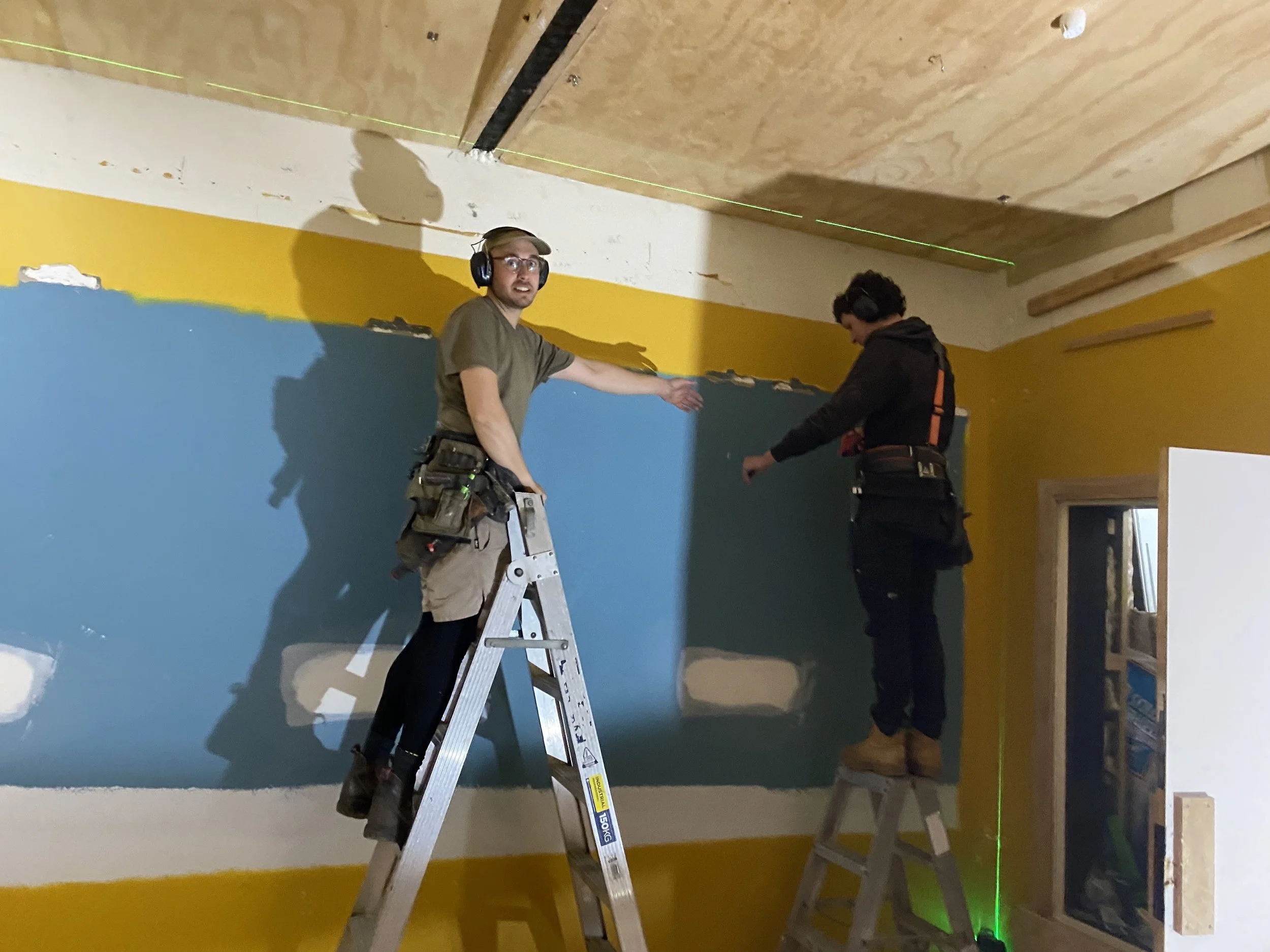 Two men wearing safety headphones working on a wall, with one standing on a ladder and the other on the ground, in a room with yellow and blue painted walls and a wooden ceiling.