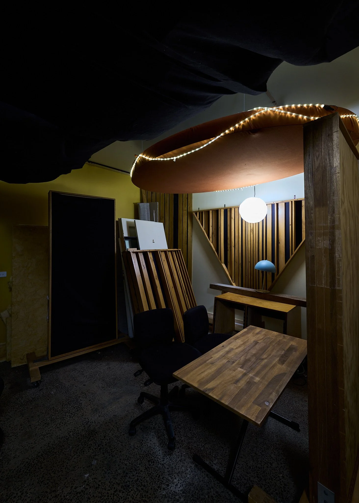 An indoor space with a wooden table, black office chairs, and shelves with wooden slats. There is a round hanging light and a curved white light fixture. The ceiling has large black fabric panels and a large pink circular ceiling feature with string lights.