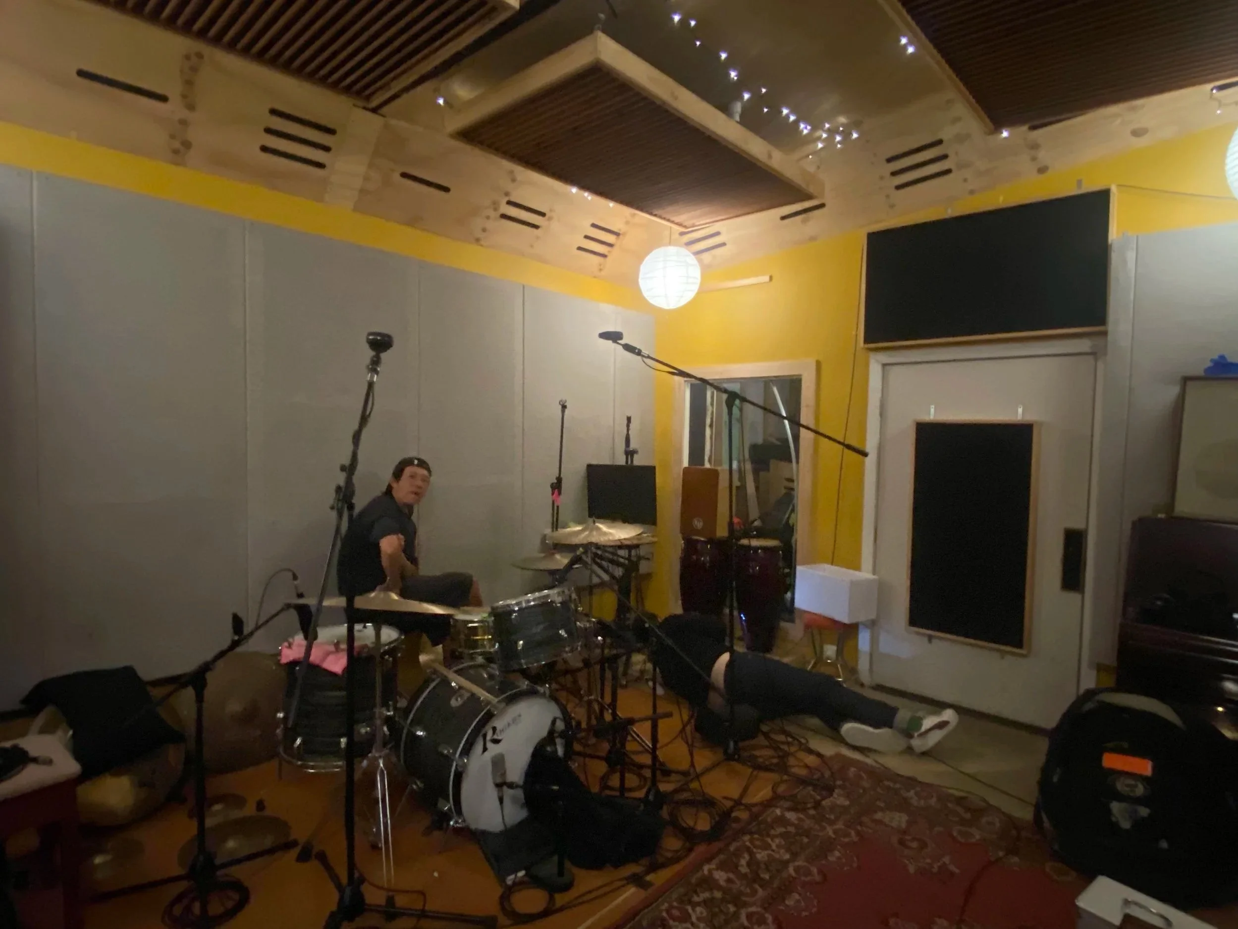 Music studio with drums, microphones, and recording equipment, yellow walls, and wood panel ceiling with string lights.
