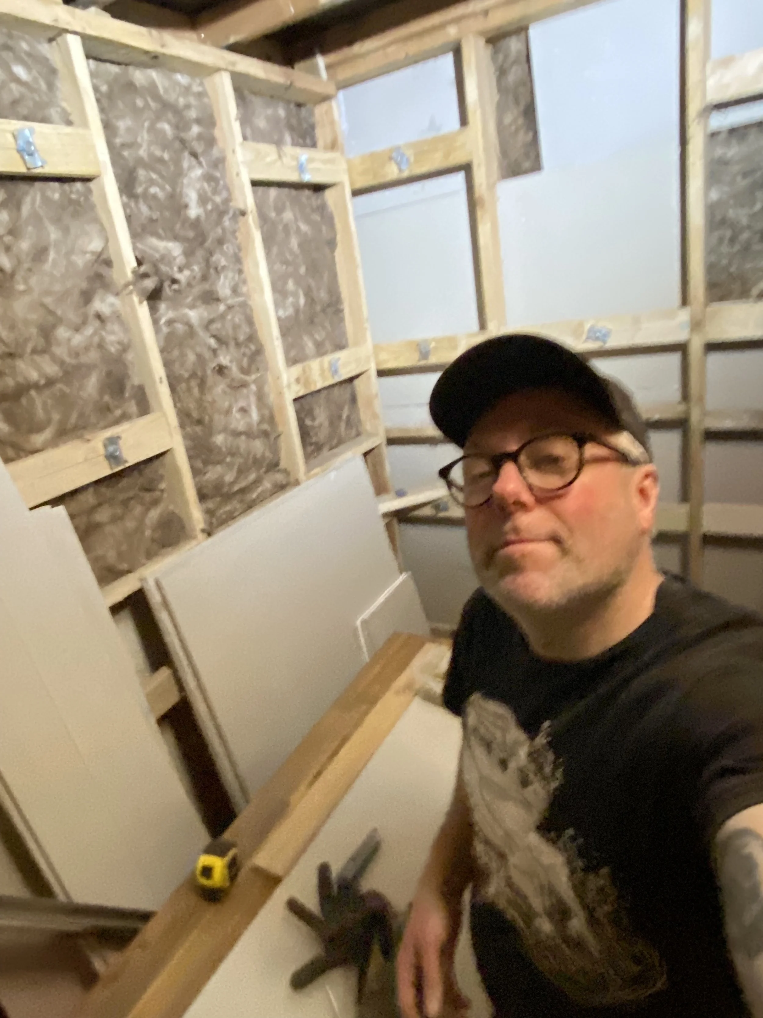 A man wearing glasses and a black cap taking a selfie in a room under construction. The wall behind him shows wooden framing and insulation panels, with some tools placed on the wooden surface.