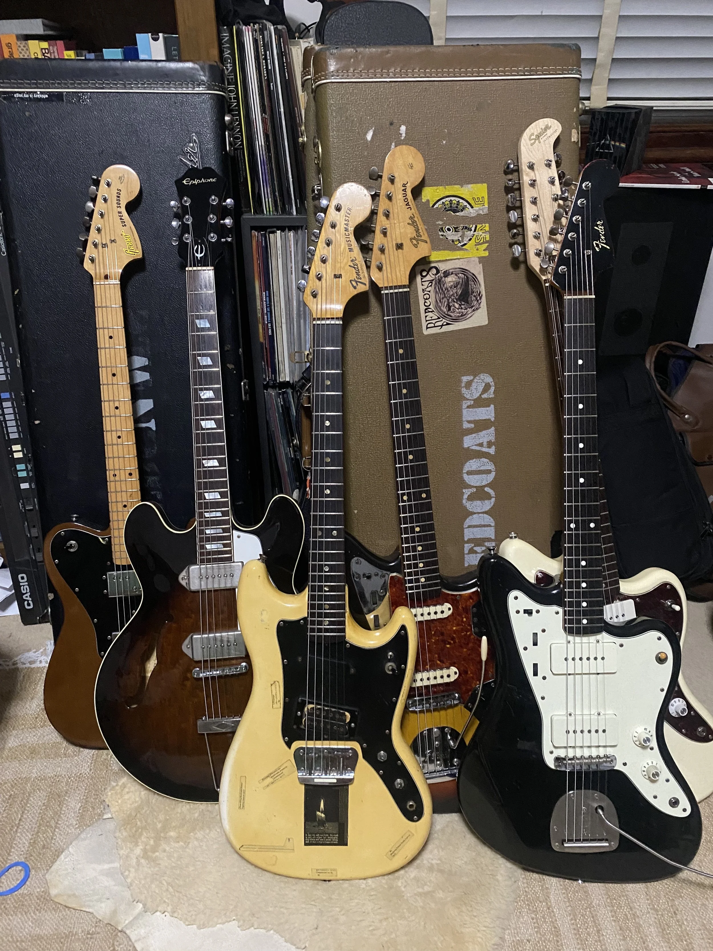Collection of six electric guitars leaning against a brown amplifier, with books and vinyl records in the background.