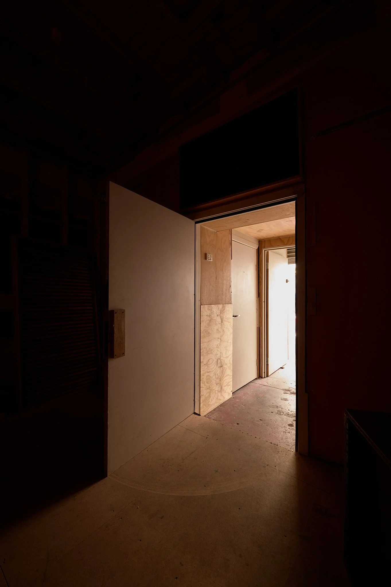 A door partially open revealing a weathered, unfinished room with wood paneling and concrete floor, lit by natural light.