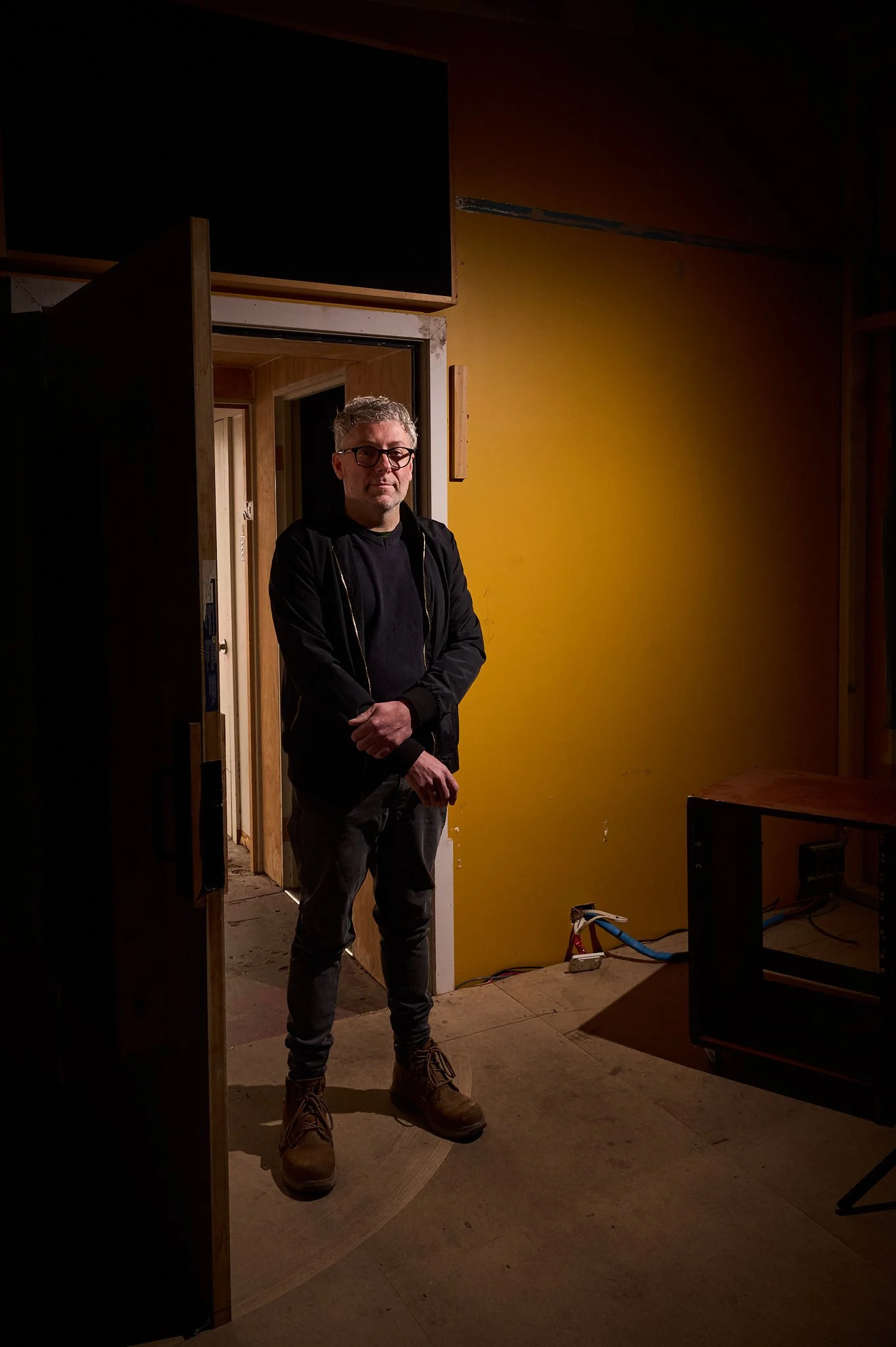 A man with gray hair, glasses, and casual clothing stands in a dimly lit room with yellow and wooden walls, near an open doorway, with construction or remodeling materials visible on the floor.