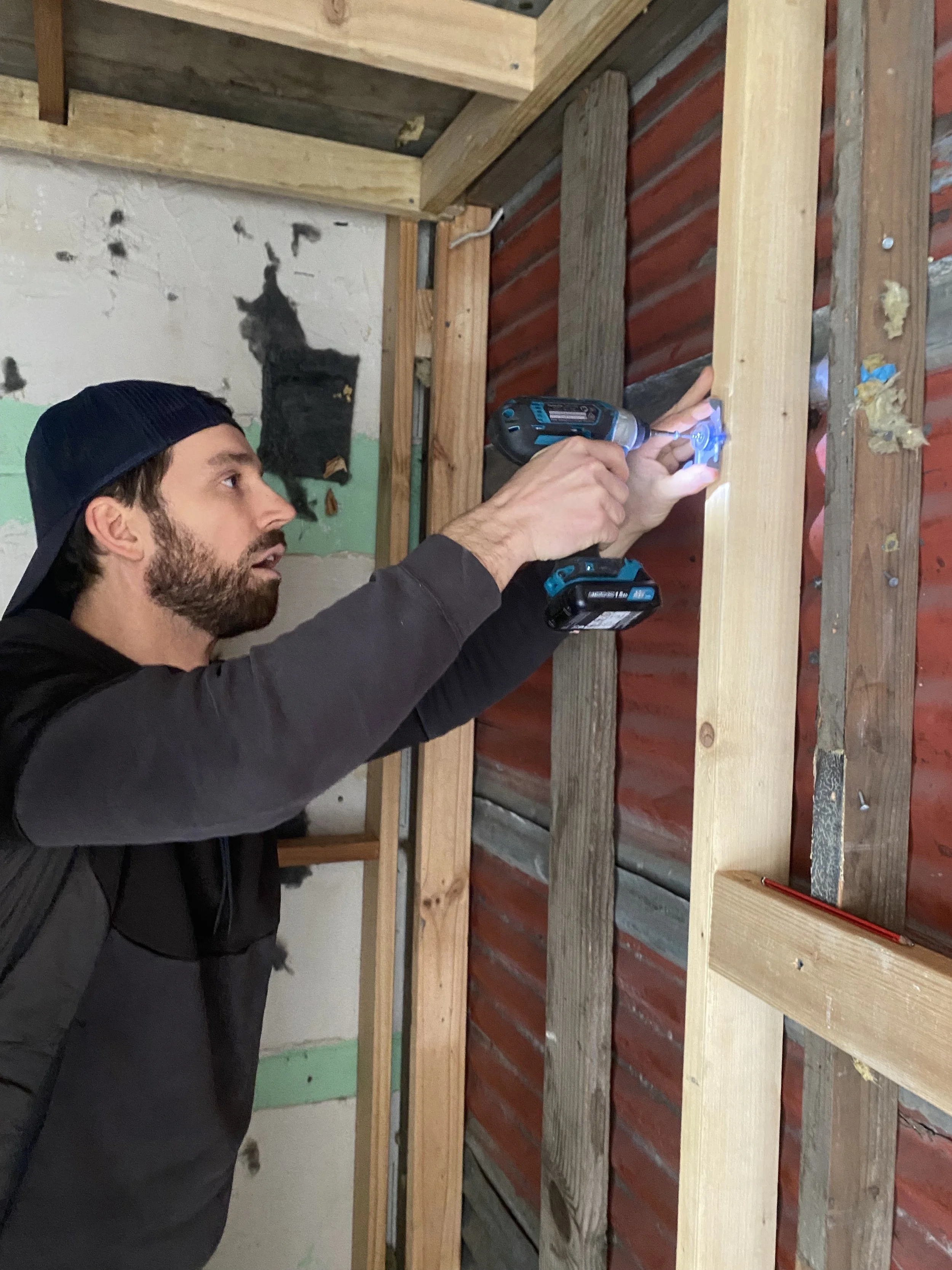 A man wearing a black cap and dark hoodie is using a cordless drill to work on a wooden frame structure at a construction site. The wall behind him has exposed wooden studs and some red-painted panels.