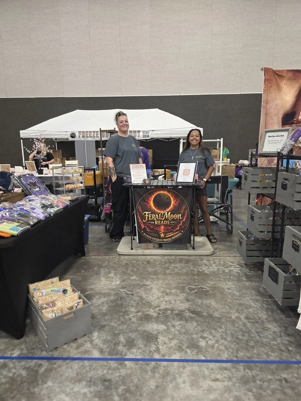 Two women standing behind a booth at a convention or fair, promoting Feral Moon Reads, a women veteran owned business. The booth has a sign with a solar eclipse graphic and the business name. Books and merchandise are displayed on tables around them.