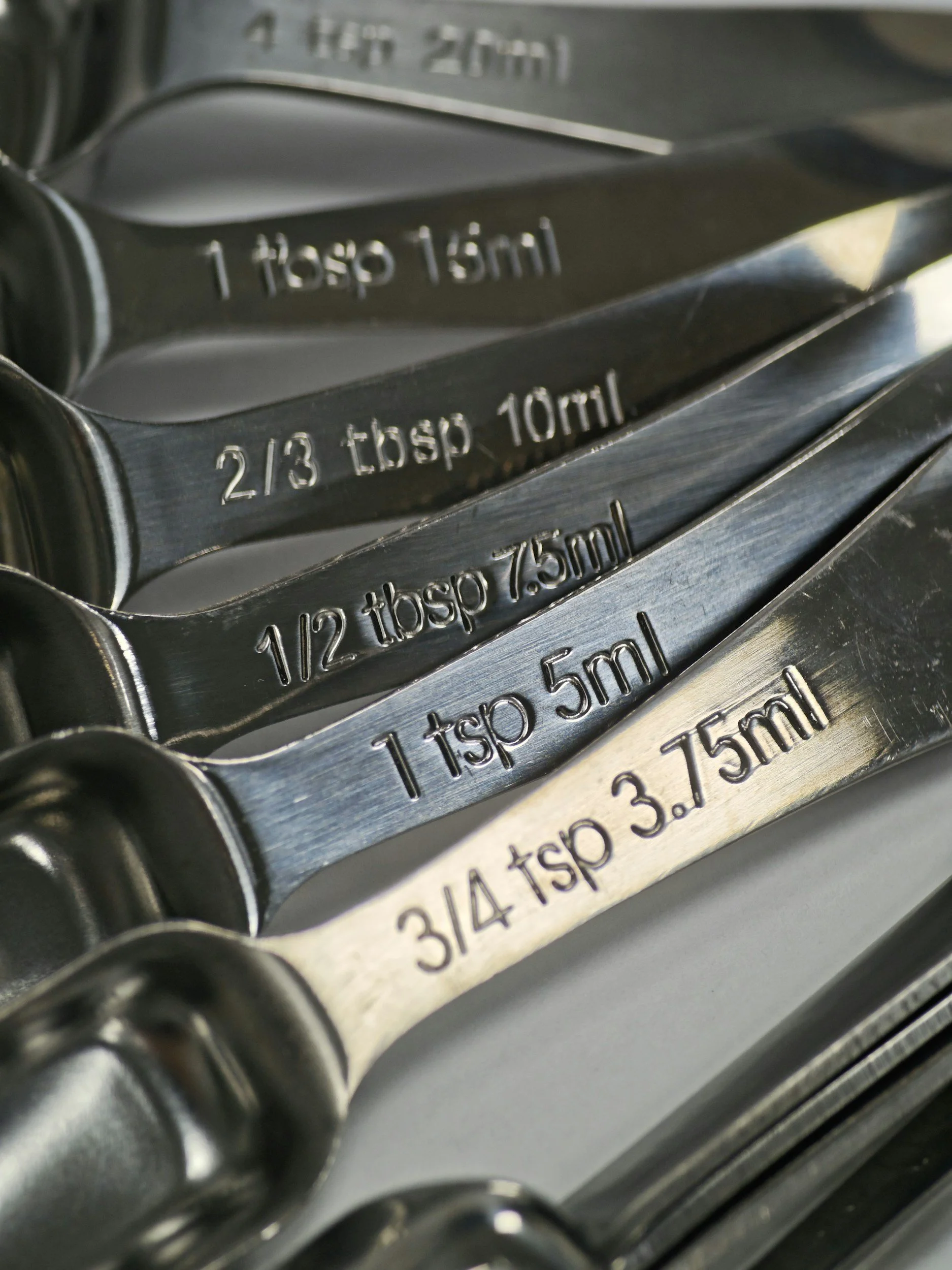 Close-up of metal measuring spoons stacked together with weight and volume measurements engraved on each spoon, including 1 tablespoon (15 ml), 2/3 teaspoon (10 ml), and 1/2 teaspoon (7.5 ml).