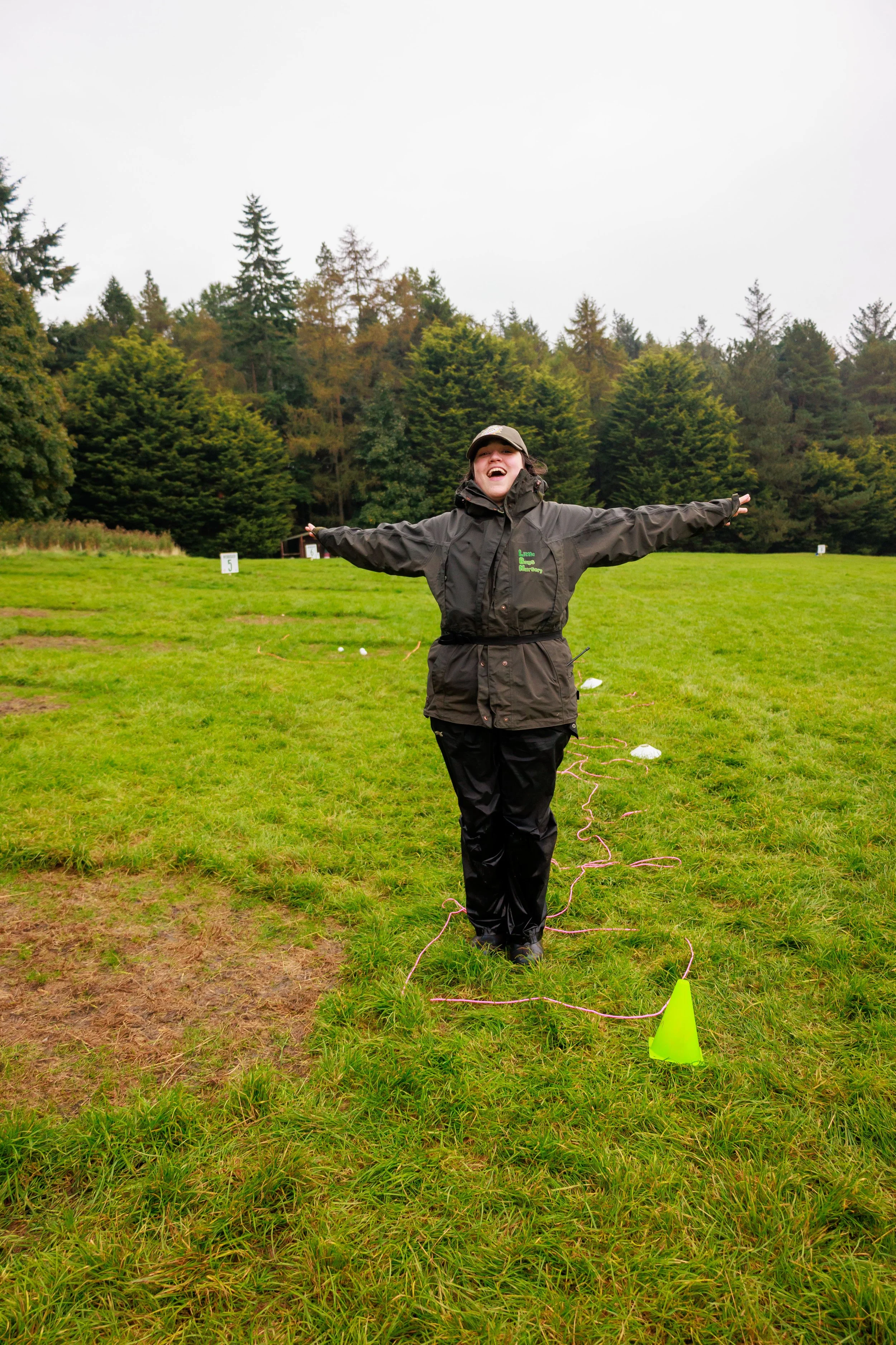 A person in a dark rain jacket and cap stands on a grassy field with arms outstretched and a big smile. The field is marked with small white signs, and a yellow cone is in the foreground. Trees line the background under a cloudy sky.