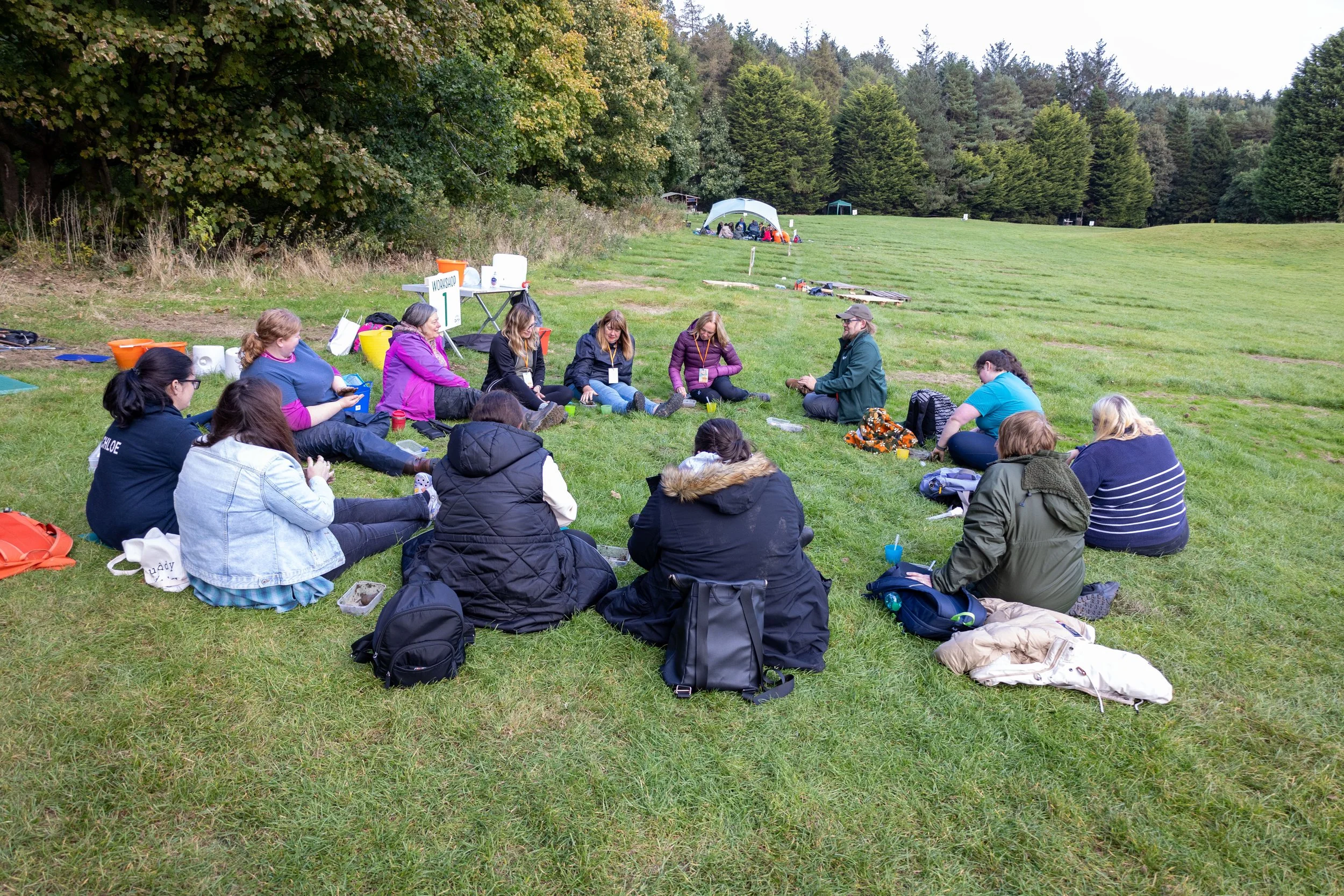 A group of people sitting on the grass in a circle outdoors, with trees and open field in the background. Some are wearing jackets and appear to be engaged in a discussion or activity.