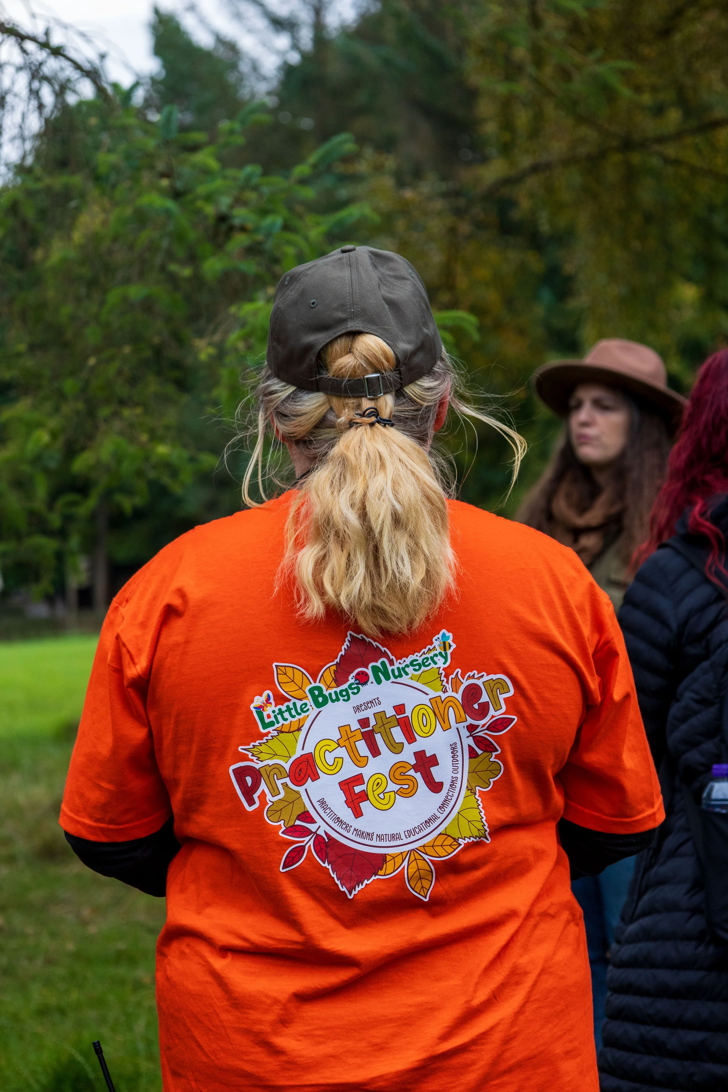Back view of a person wearing an orange T-shirt with a logo that says 'Little Bugs Nursery' and 'Practitioner Fest' with people gathered outdoors in a park or garden with green trees.