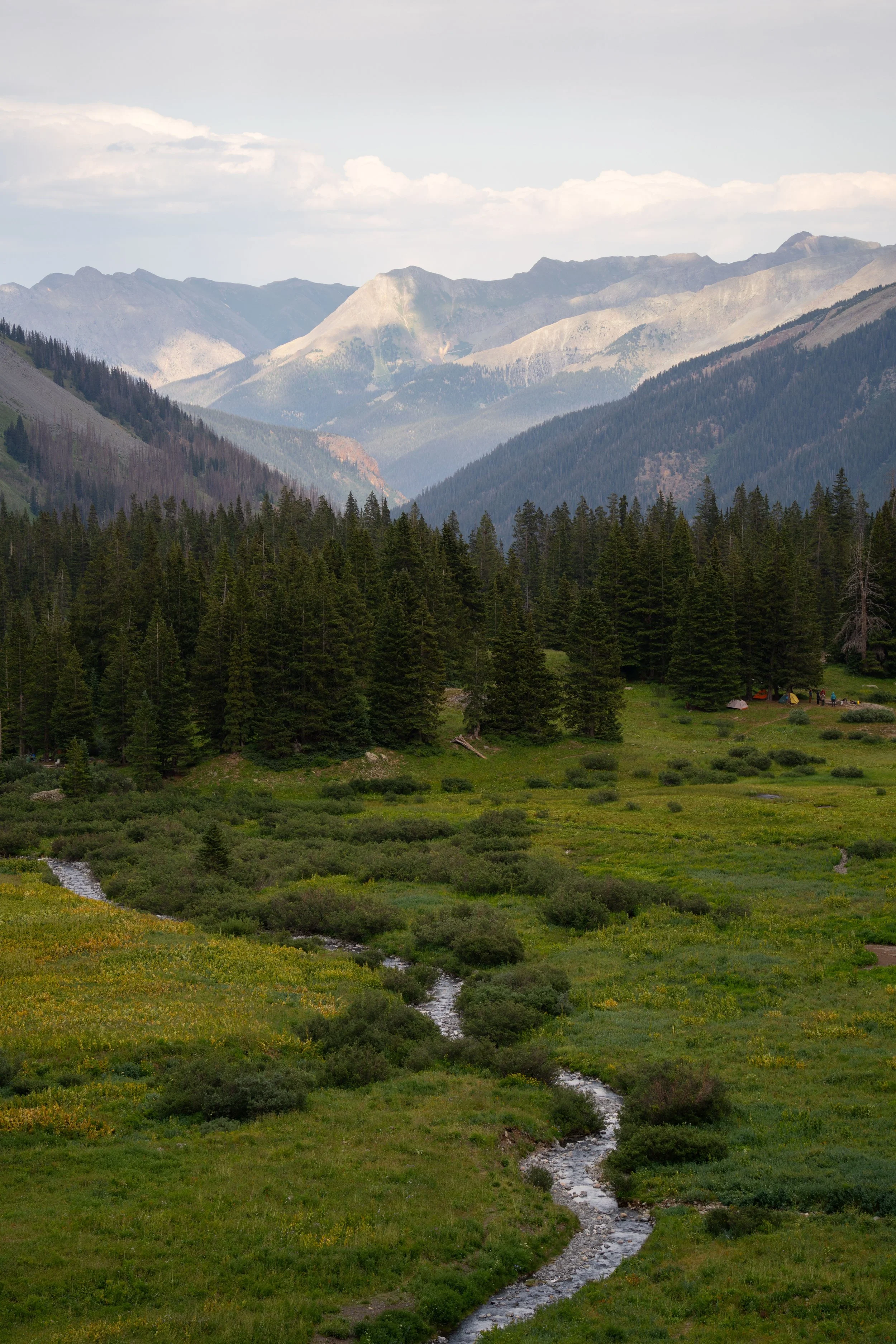 Scenic view of a mountainous landscape with a lush green valley and a small winding stream in the foreground, dense forest, and tall mountain peaks in the background, under a partly cloudy sky.