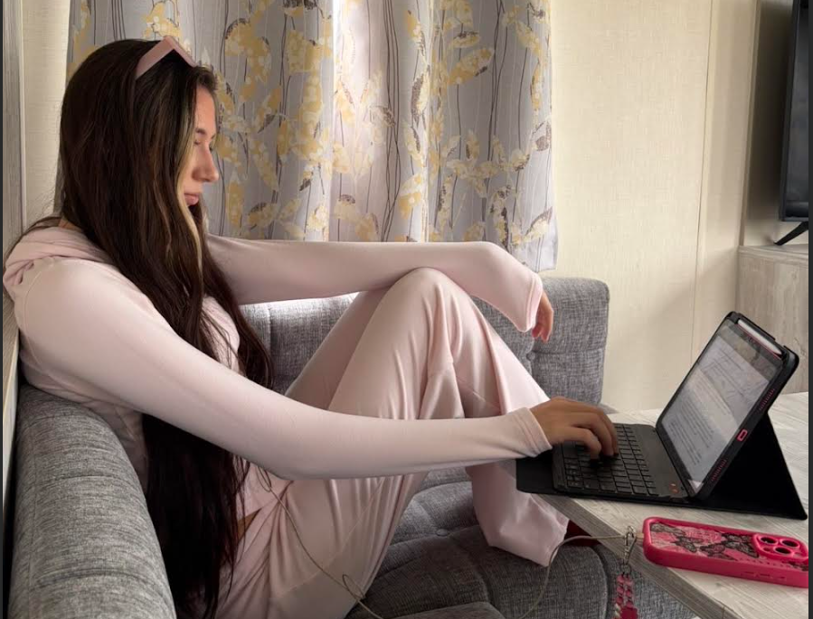 A young woman with long brown hair, wearing pajamas, sitting on a gray couch with her legs bent, working on a laptop at a small table in a living room. There are curtains with a floral pattern in the background and a pink phone on the table.