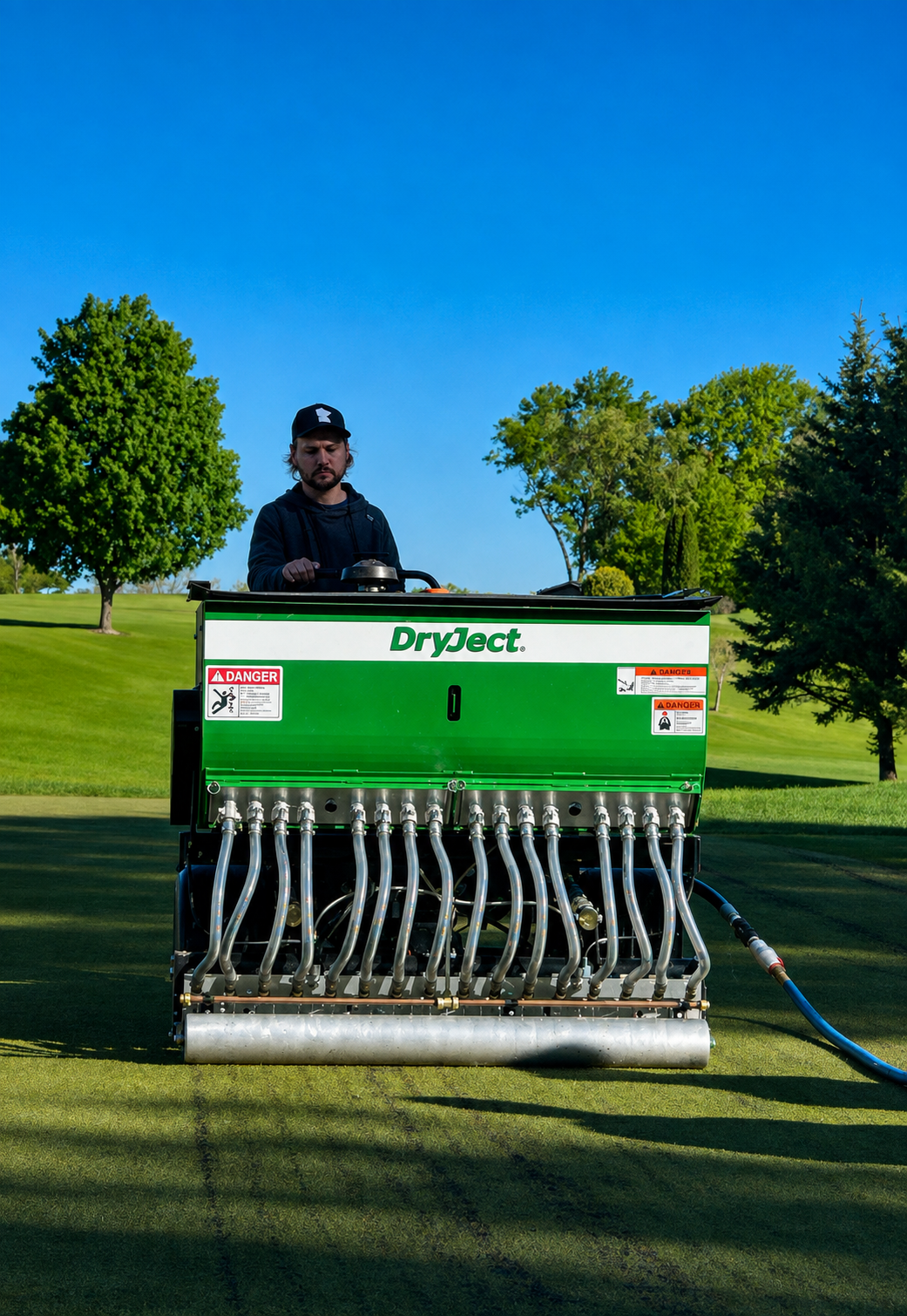 A man is operating a green DryJect machine on a golf course, watering the grass with a blue hose attached to the machine. The scene is outdoors with trees and clear blue sky in the background.