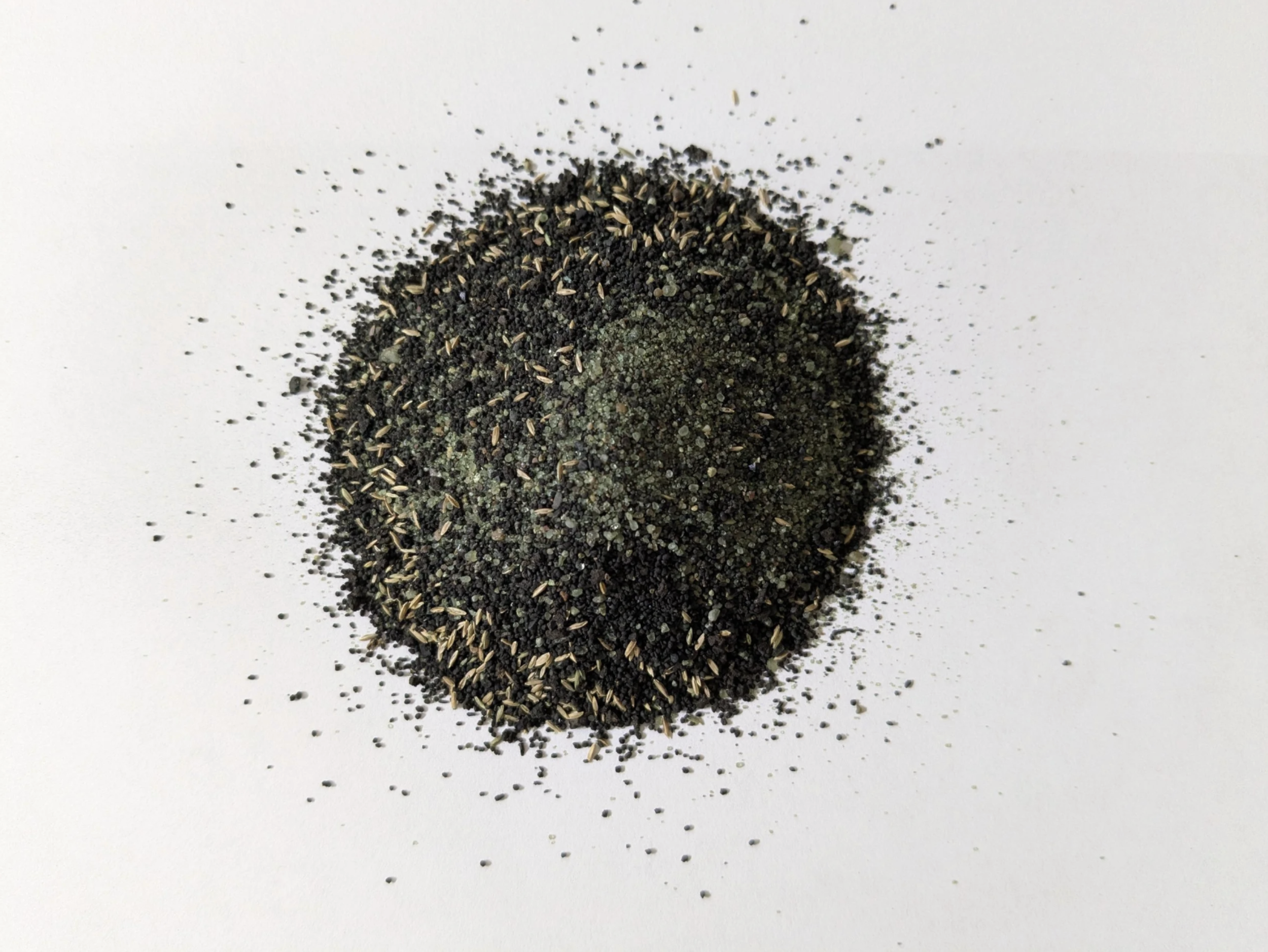 A mixture of black and greenish-brown seeds or herbs spread on a white surface.