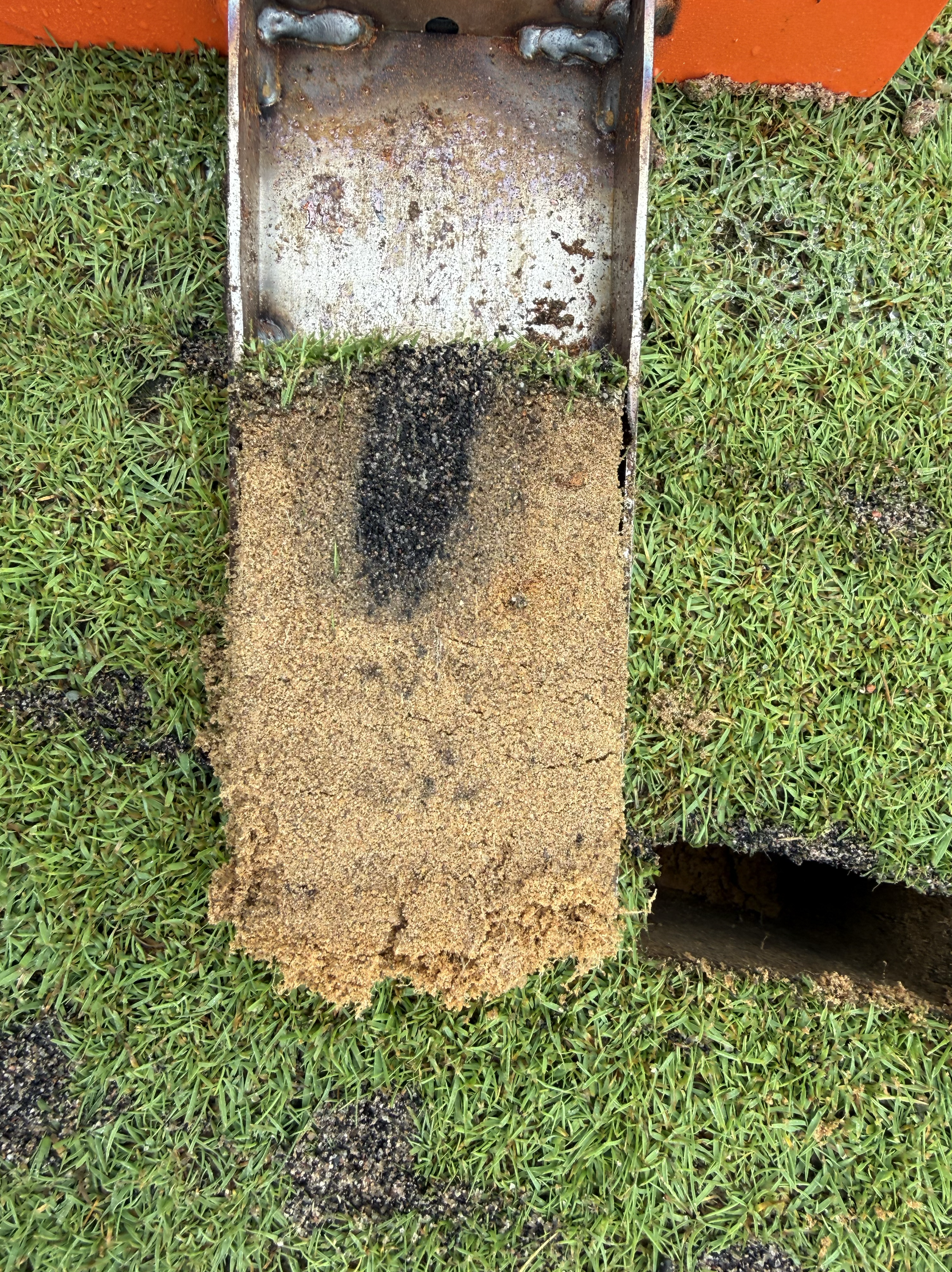 A metal U-shaped bracket holding a concrete slab in place, surrounded by green grass, with a hole underneath the slab revealing dirt.