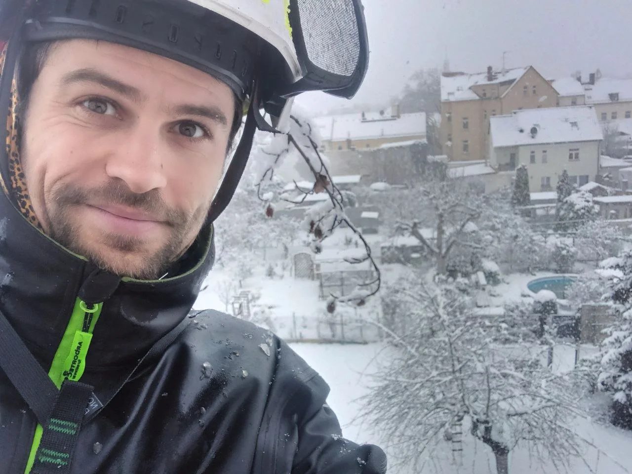 A man wearing a black helmet and jacket stands outside in a snowy landscape during daytime, with snow-covered houses and trees in the background.