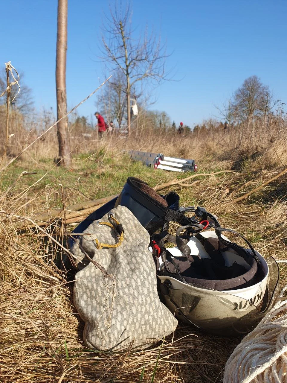 A black and white bicycle helmet, a patterned gray pouch, and a device are on dry grass in a natural outdoor setting on a sunny day. In the background, there are leafless trees, a few people walking, and a ladder on the ground.