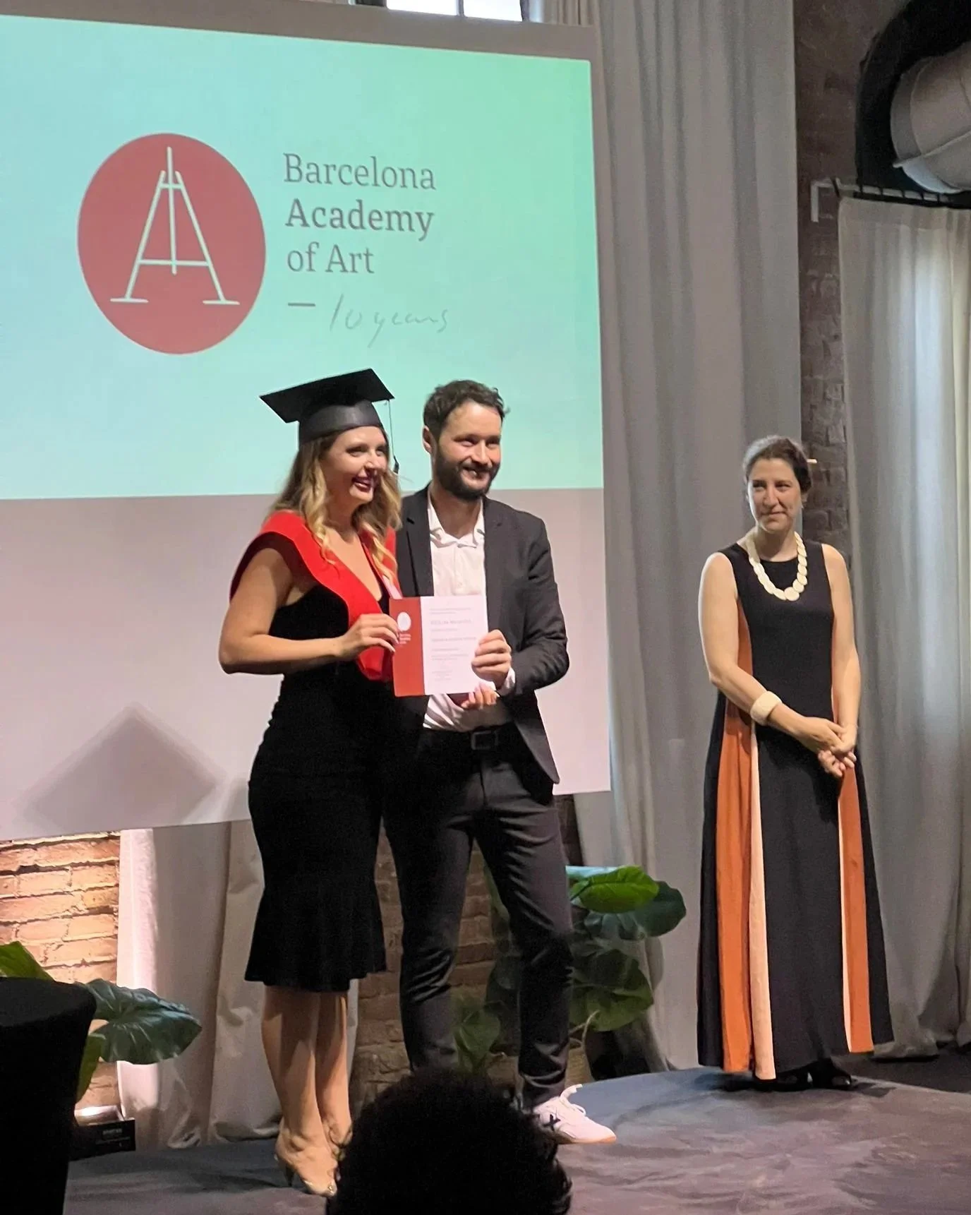 A woman in a black dress and graduation cap is holding a diploma and smiling at an award ceremony. A man in a suit is standing beside her, smiling. A woman in a long black and orange dress stands to the right, watching. A large screen behind them displays the logo and name of the Barcelona Academy of Art with the text indicating a 10-year celebration.