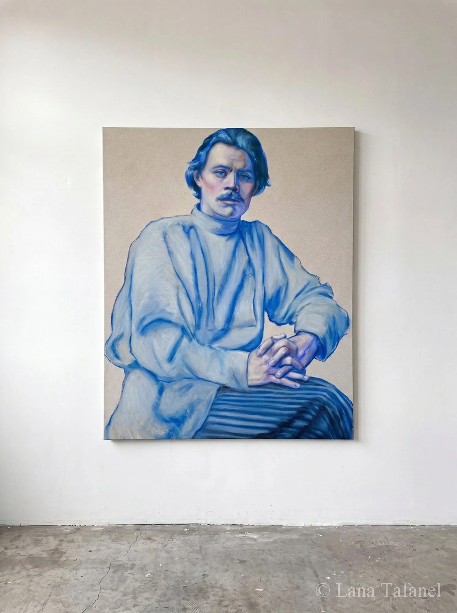 A large portrait painting of a man with hair, a mustache, and beard, seated with his hands clasped. The man is wearing a light-colored shirt, and the background is neutral. The painting is displayed on a white wall in an art gallery or studio space w