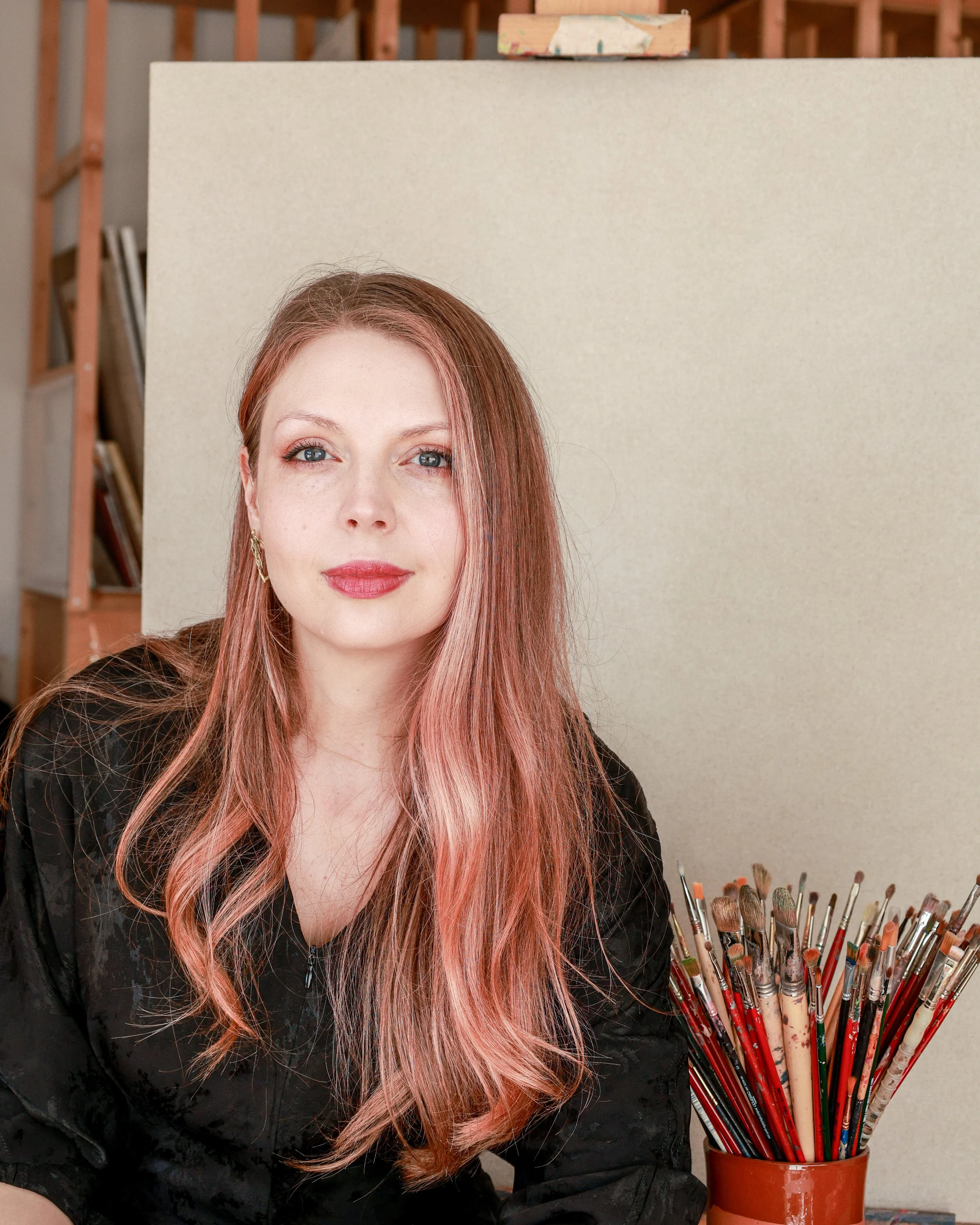 Portrait of Lana Tafanel, painter, looking at the camera in her studio with an easel and brushes in the background.