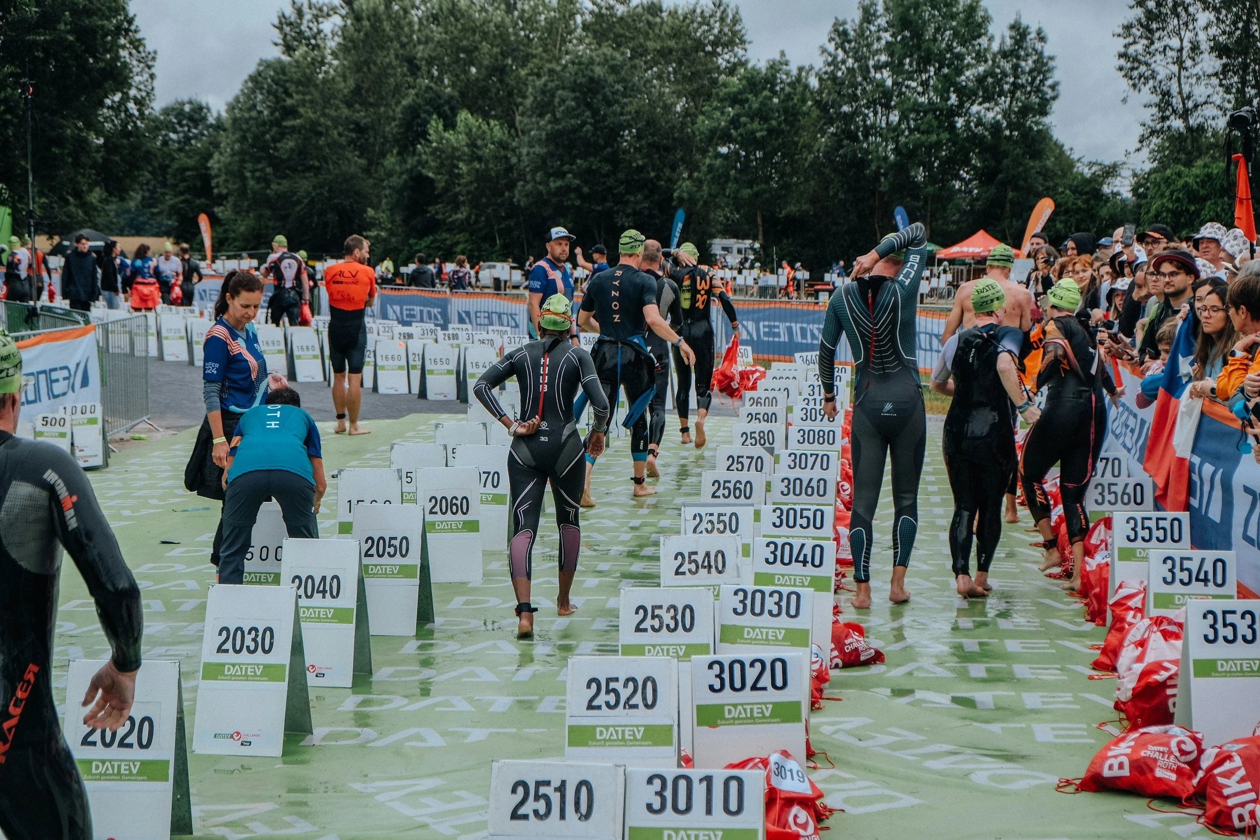 Triathlon transition area with athletes in wetsuits, green swim caps, and numbered bike and run gear, surrounded by spectators and event banners on a cloudy day.