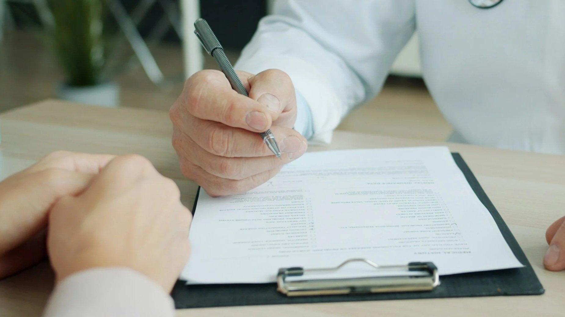 A person in a white coat signing a document on a clipboard with a pen, while another person's hand is visible on the table.