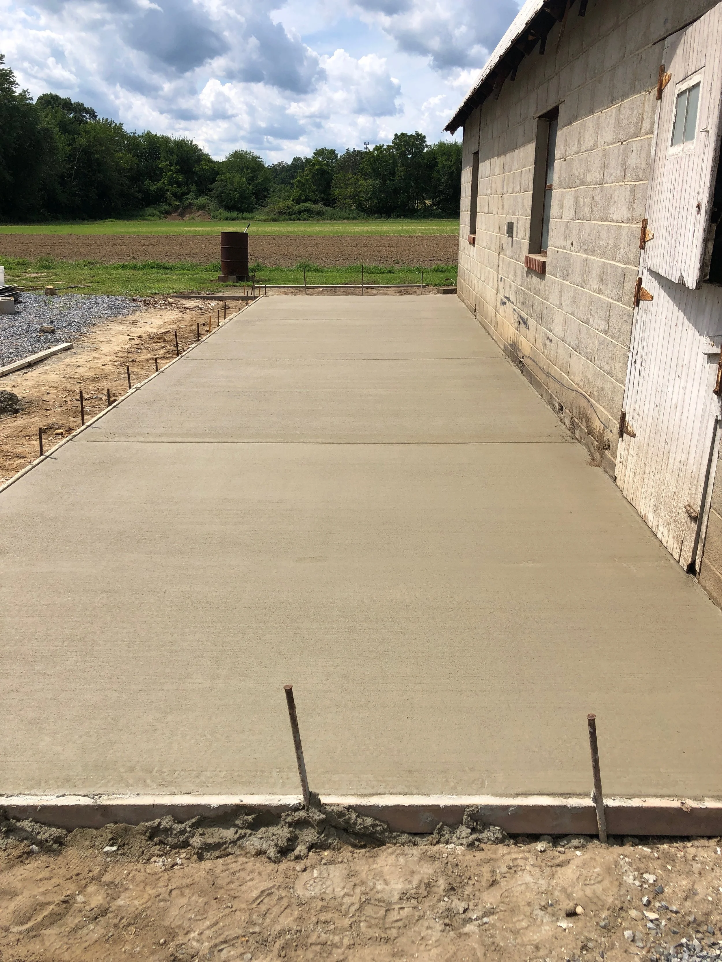 Newly poured concrete slab foundation for a building, with a partially constructed wall on the right and a field and trees in the background.