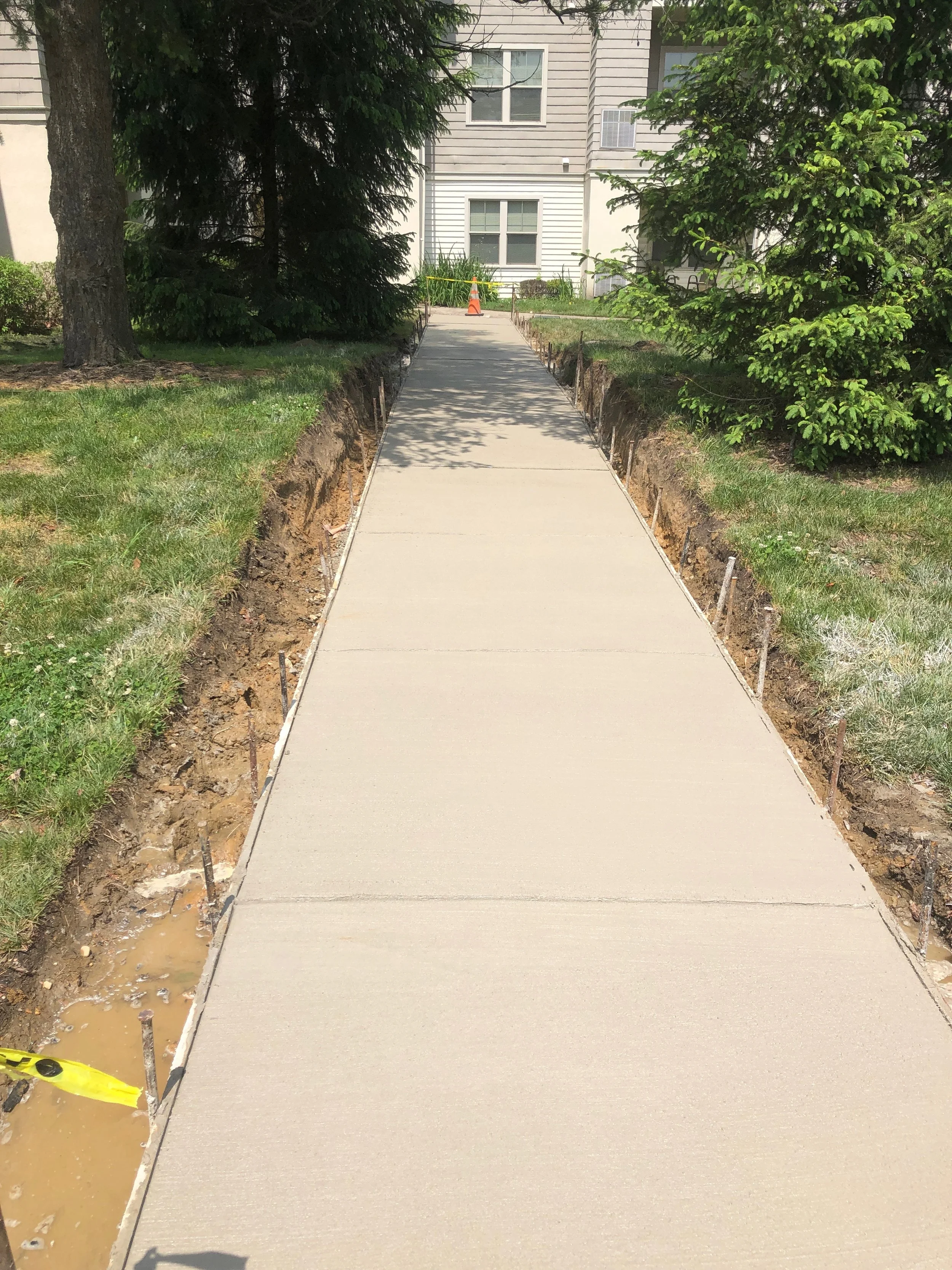 Newly poured concrete sidewalk under construction in front of residential buildings, with trees casting shadows and a traffic cone in the background.