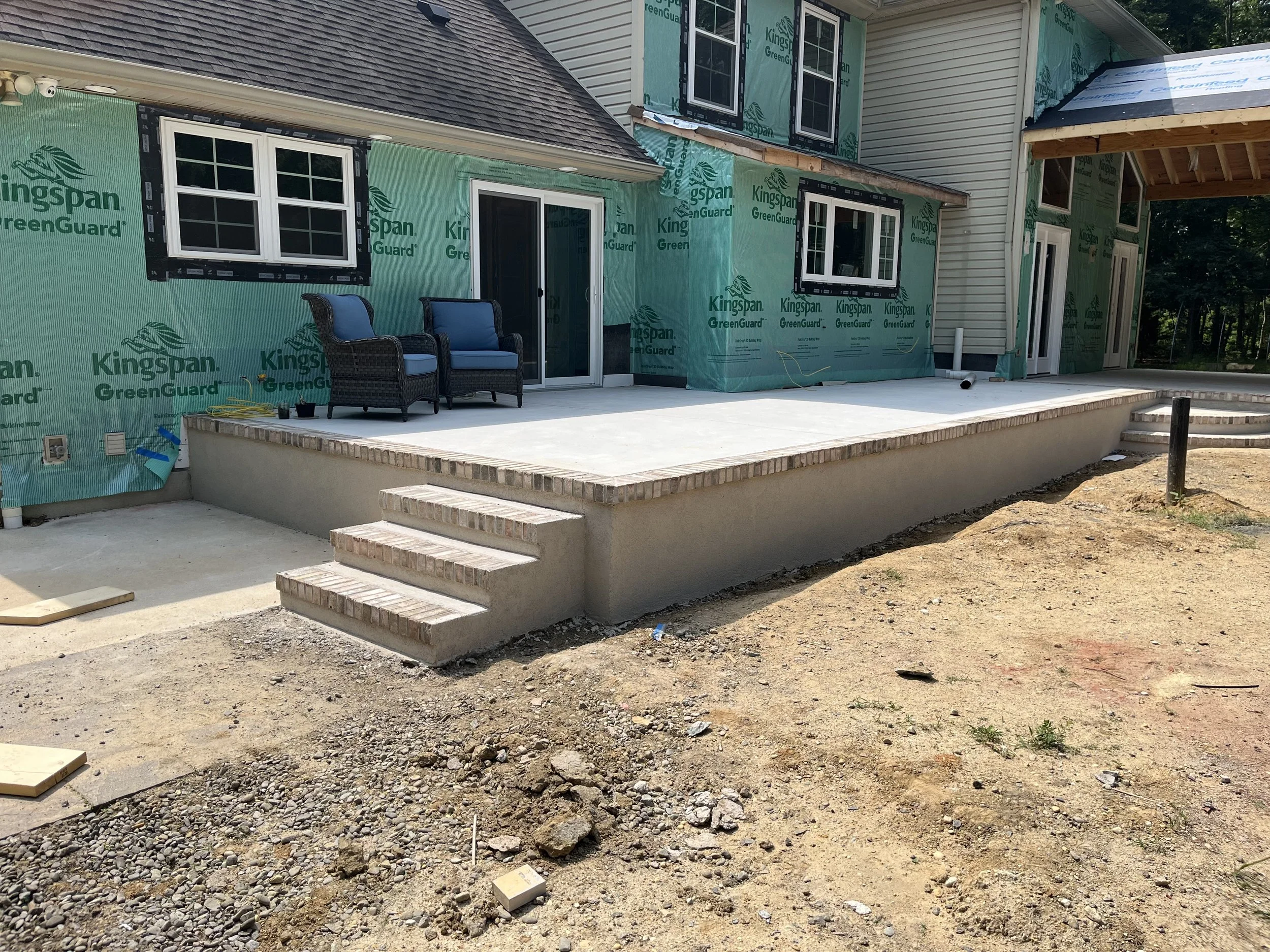 Exterior of a house under construction with green house wrap, multiple windows, a sliding door, and a patio area with outdoor chairs. Steps leading up to the patio are visible, with construction materials and dirt in the foreground.