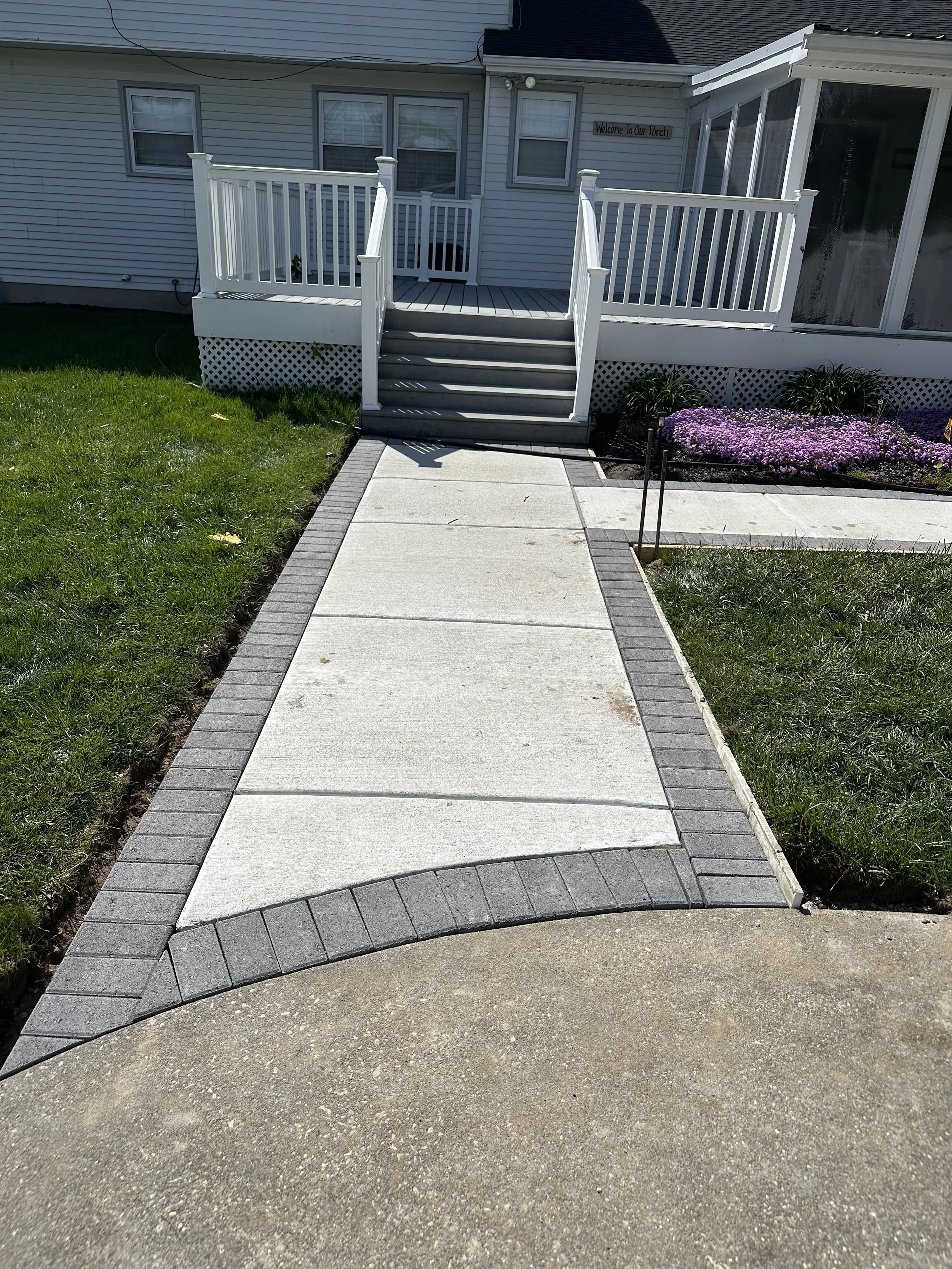 A backyard with a concrete sidewalk leading to a small set of steps up to a white deck with railings attached to a house. The house has light gray siding and multiple windows. There are purple flowers and some greenery in flower beds along the house.