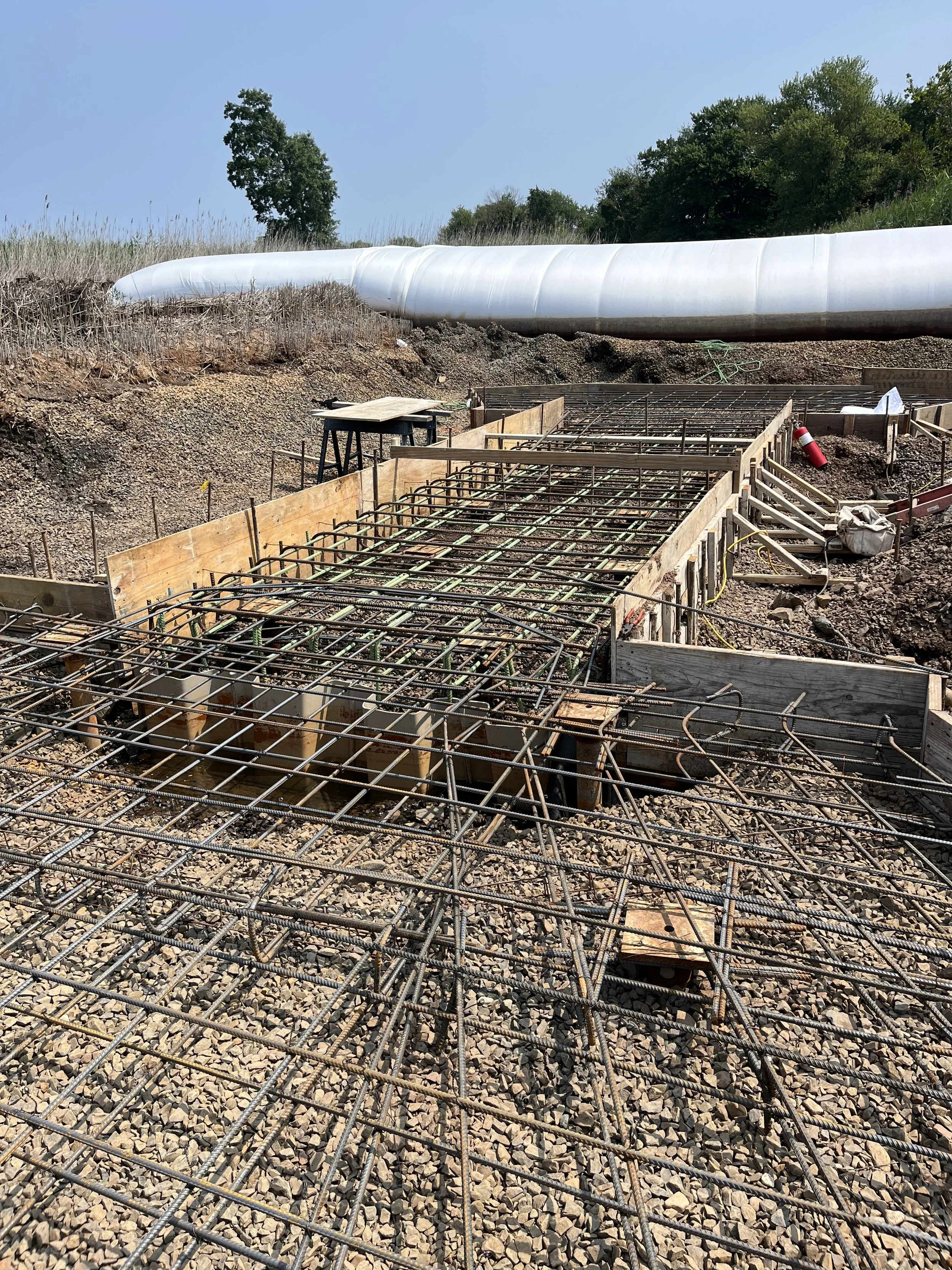 Construction site with rebar framework for concrete pouring, a fire extinguisher, and a large white pipe in the background against a clear blue sky with trees.