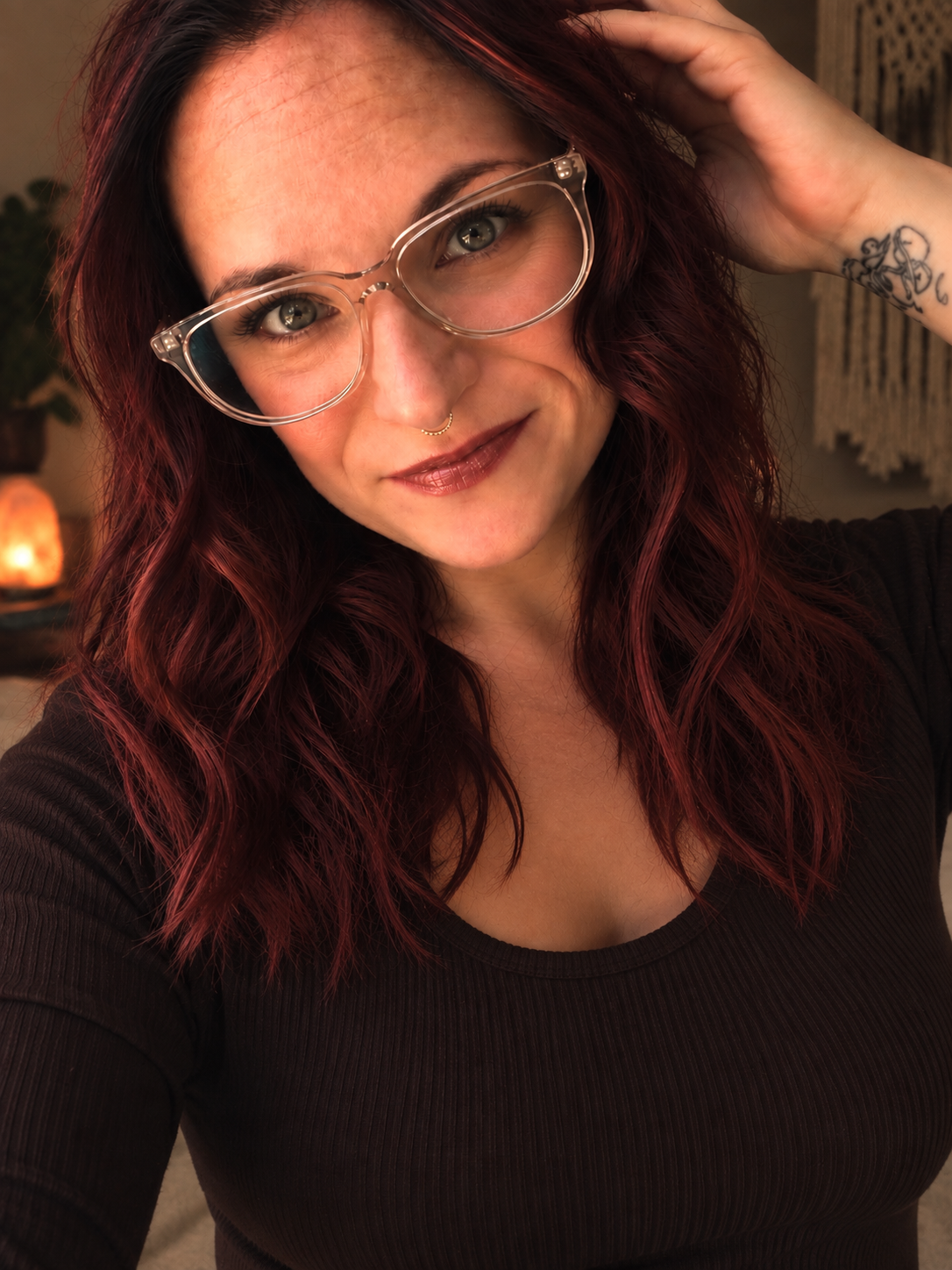 A woman with wavy red hair wearing glasses, a septum piercing, and a tattoo on her wrist, smiling and looking into the camera.