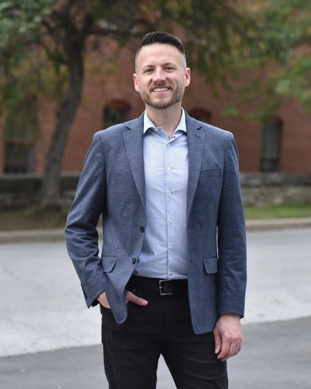 A smiling Aaron Crossley in a gray-blue blazer and light blue shirt standing outdoors in front of a brick building and trees.