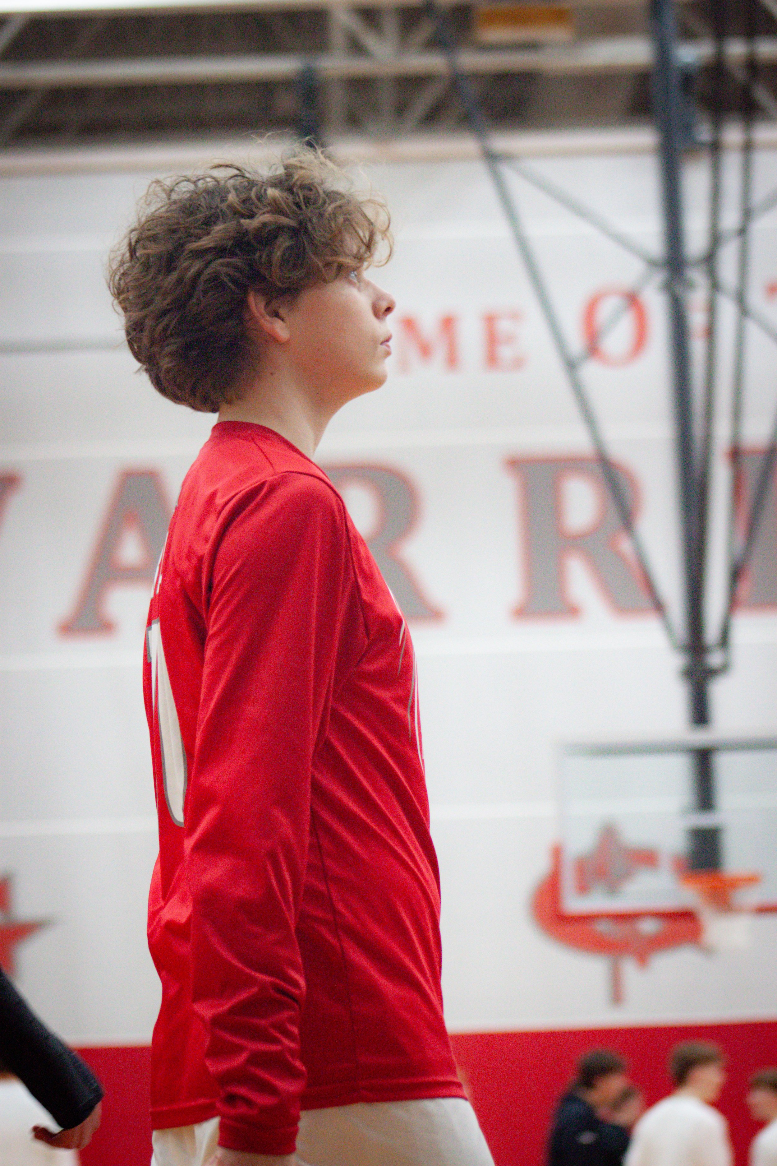 A person with curly hair wearing a red sports jersey standing in an indoor sports facility with a blurred background of spectators and equipment.