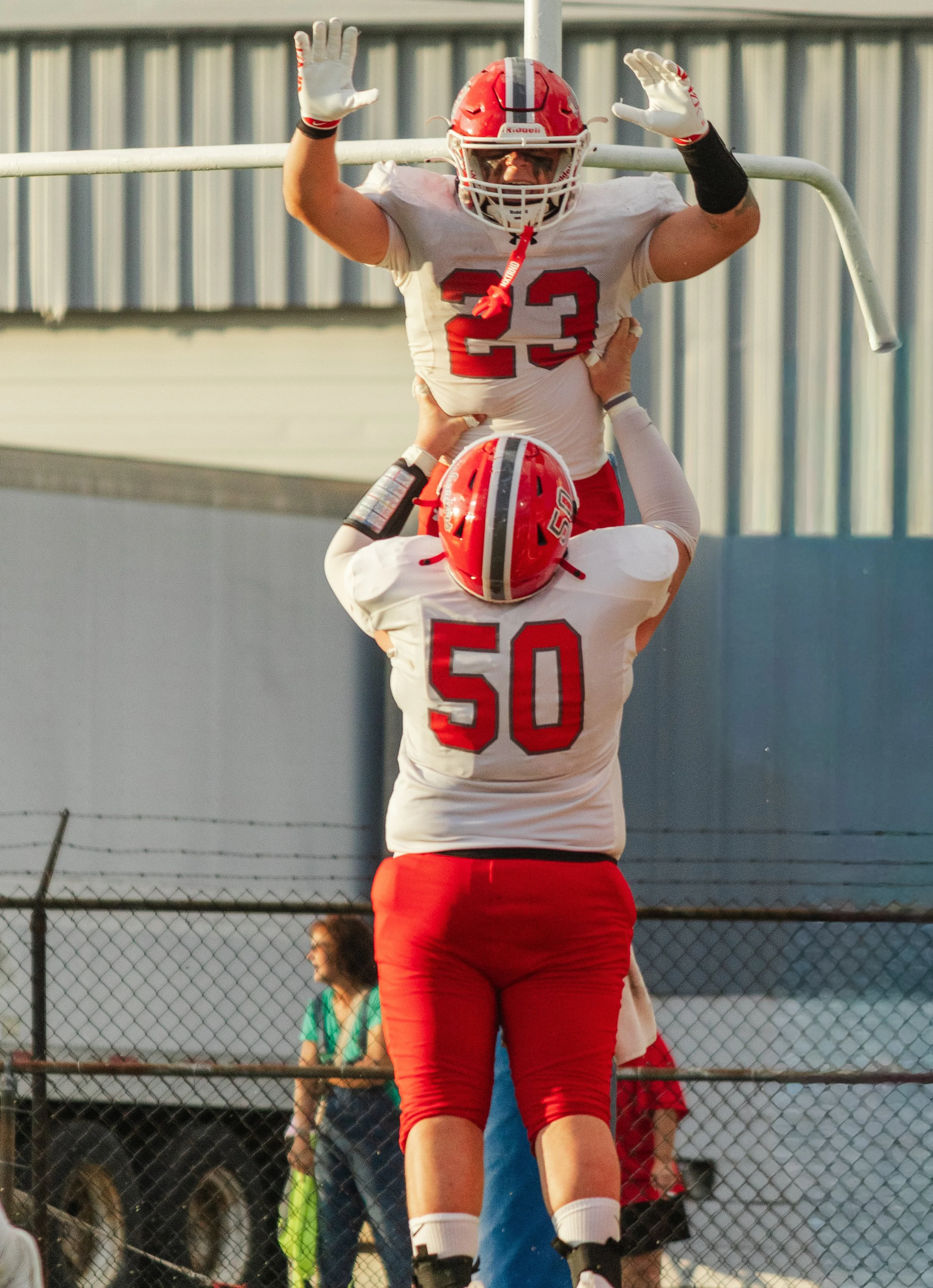 Two football players in white and red uniforms celebrating a touchdown behind a chain-link fence, with spectators behind them.