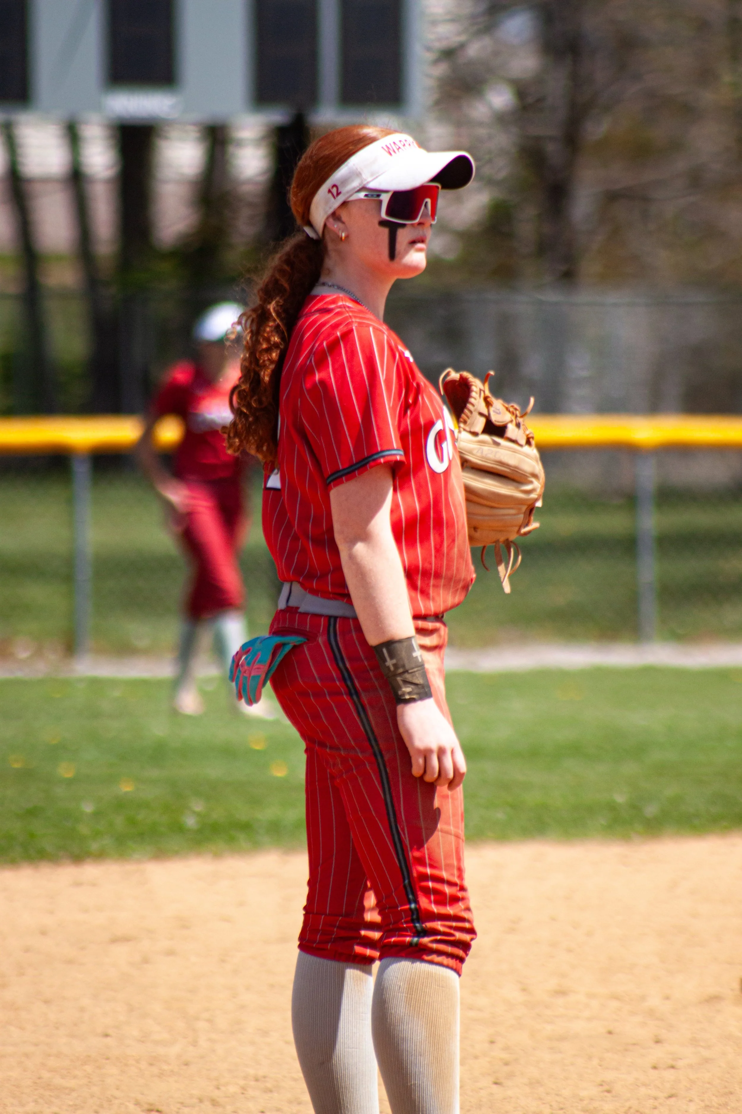 A female softball player with red hair in a ponytail, wearing a red and white striped uniform, white knee-high socks, and gray knee pads, standing on the field with a glove on her left hand. She is also wearing a white visor, red sunglasses, and ear earrings, and she appears to be focused on the game.