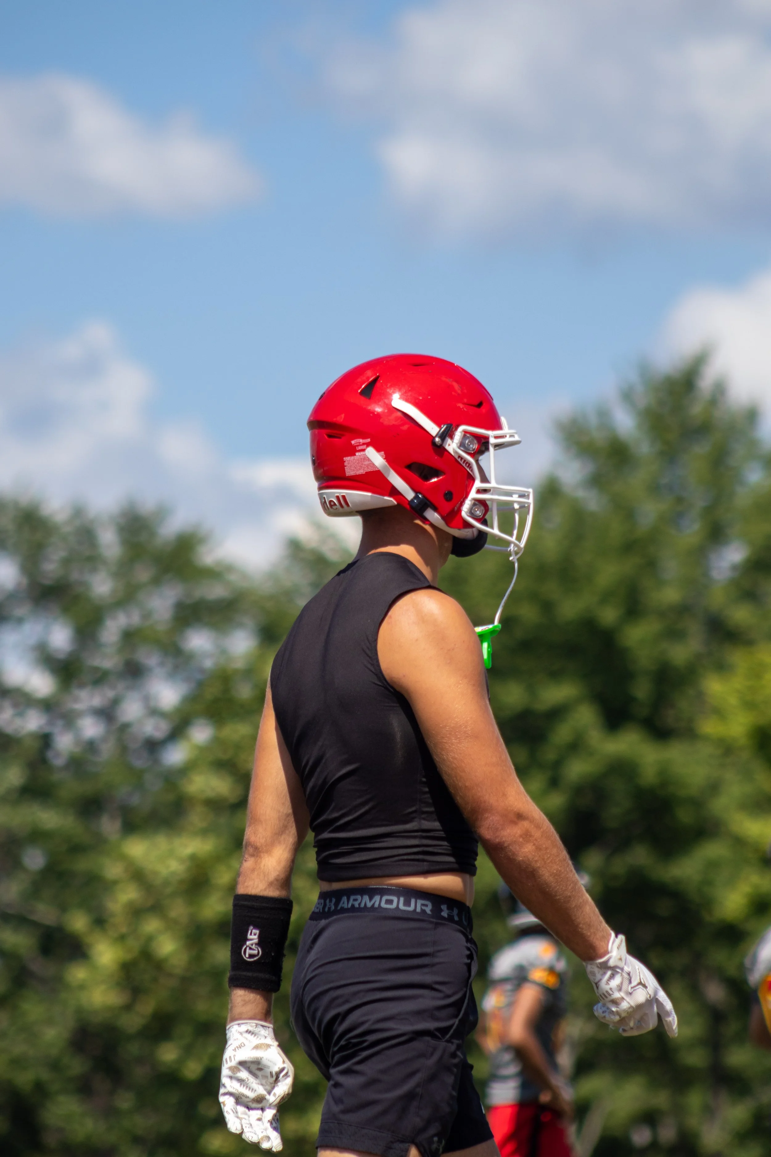 A football player wearing a red helmet, black Under Armour shirt, and black athletic shorts, standing outdoors with blue sky and green trees in the background.
