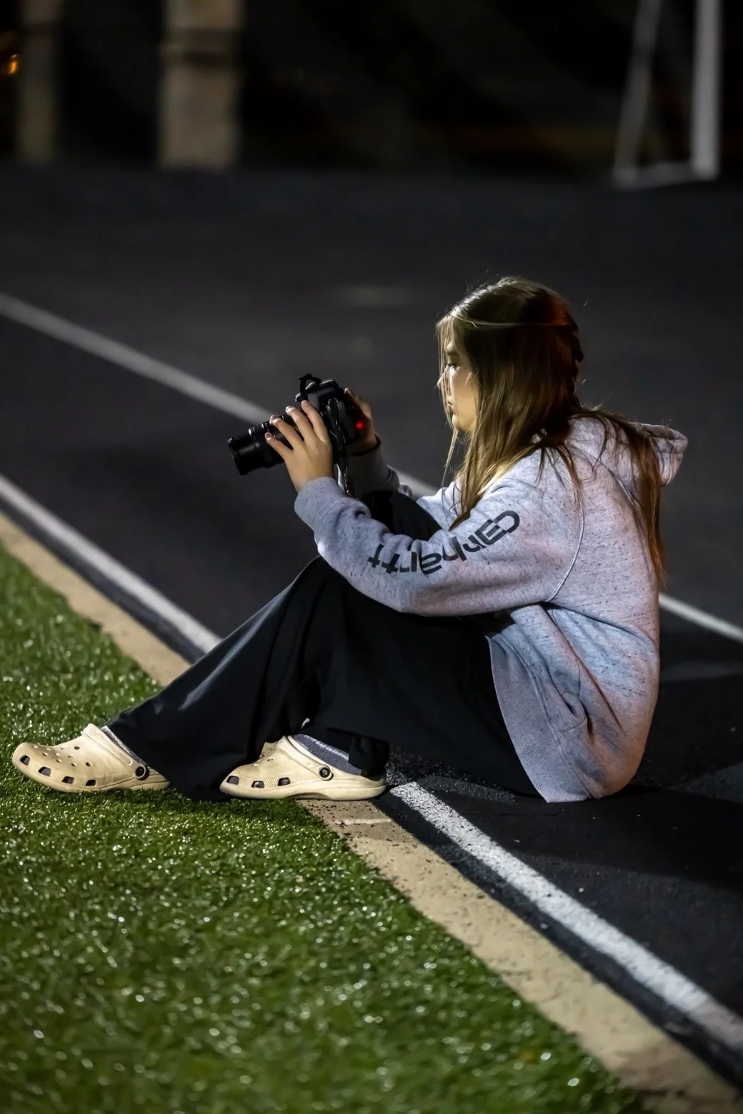 A young woman sitting on the edge of a running track at night, holding a camera and preparing to take a photo, wearing a gray hoodie and Crocs.